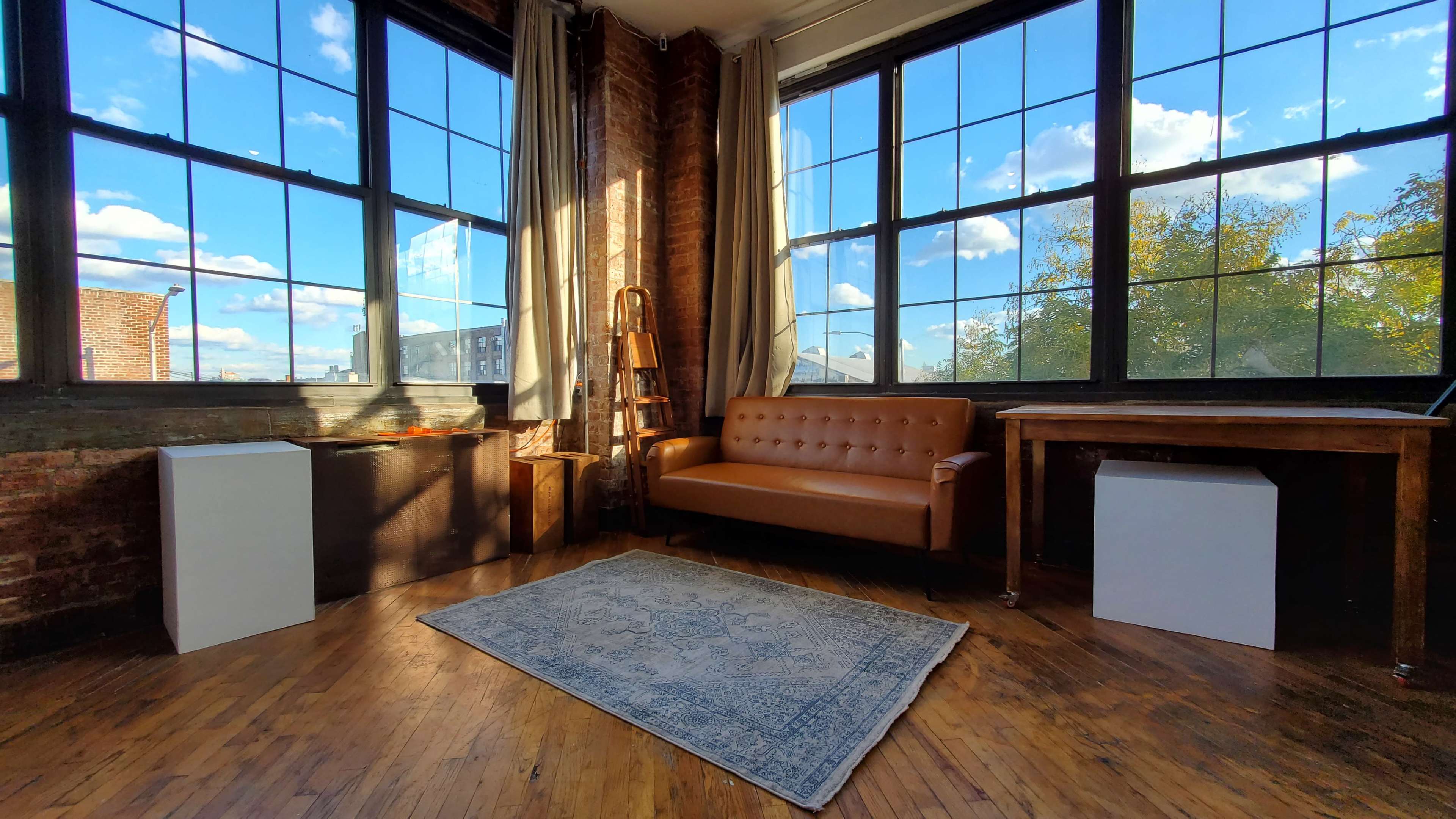 A spacious room with large windows, featuring a brown leather sofa, a wooden table, and two contrasting storage units, all illuminated by natural light.