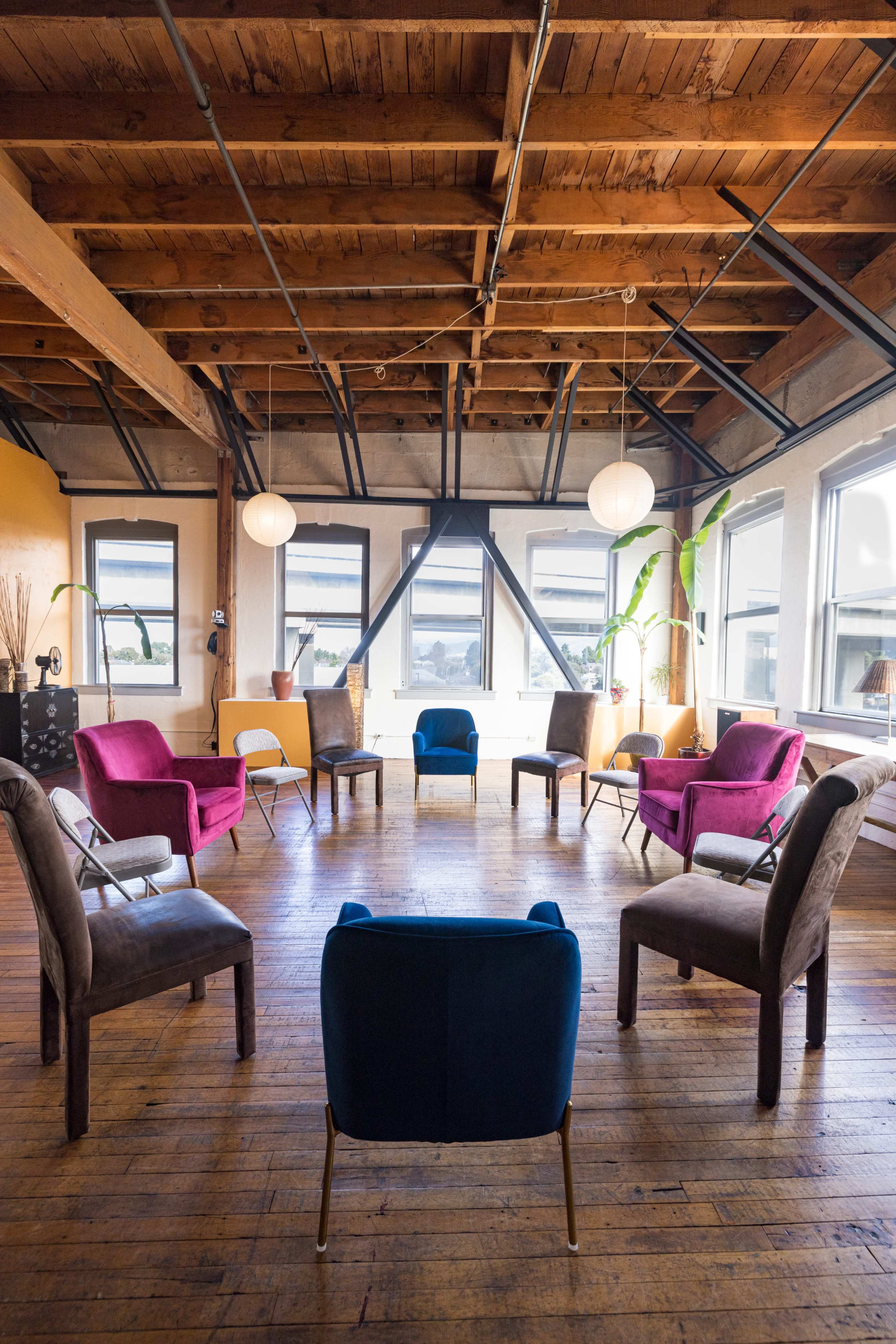 A spacious room features a circular arrangement of variously colored chairs on a wooden floor, surrounded by large windows and illuminated by pendant lights.