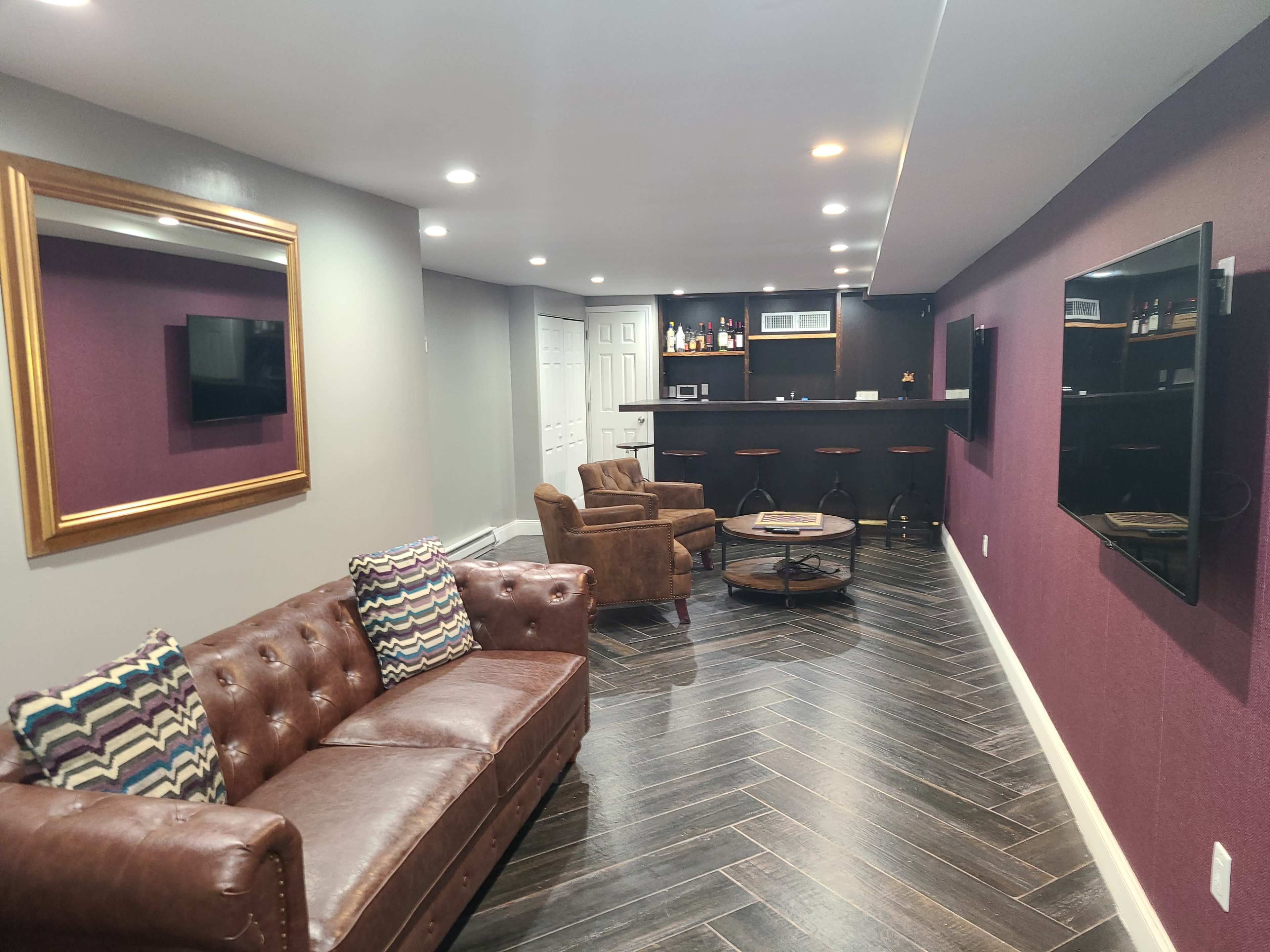 A modern basement features a leather sofa, two armchairs, a round coffee table, and a bar area with stools against a dark purple wall.
