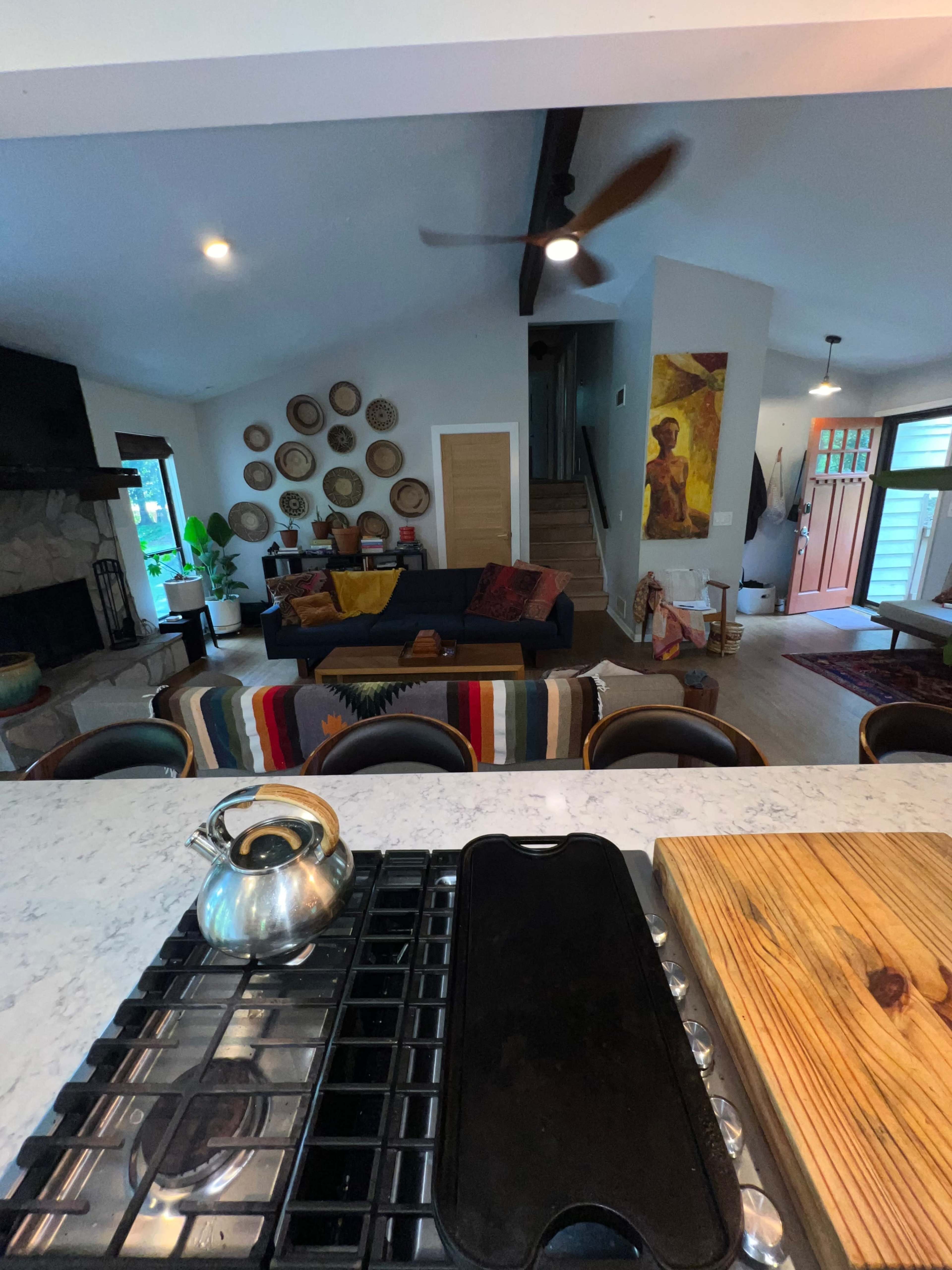 A modern living space featuring a kitchen counter with a kettle, a stove, and a view into a living room with a dark sofa and wall art.