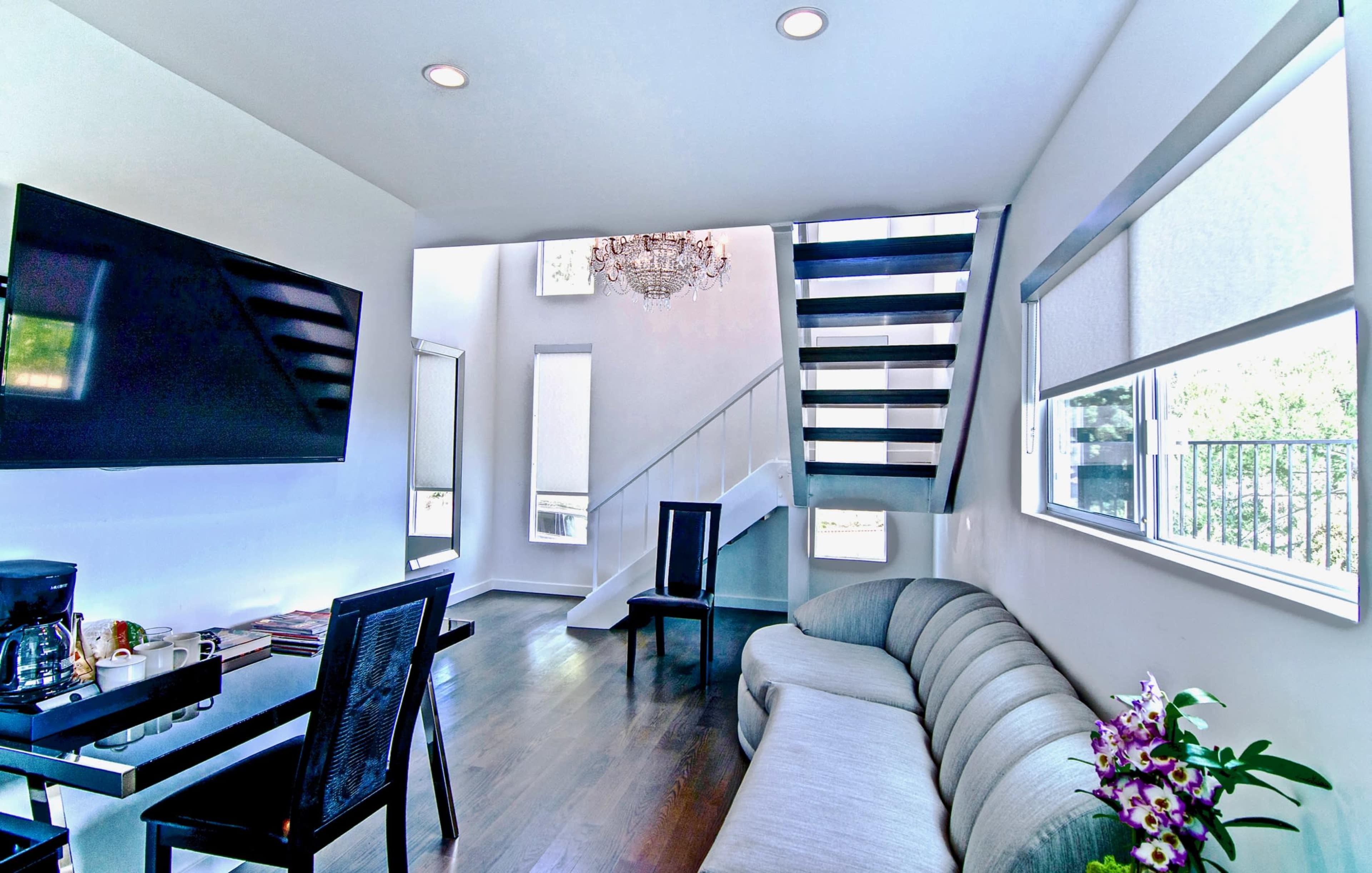 The image shows a modern living area with a gray curved sofa, a glass dining table, and a staircase leading to an upper level, all illuminated by natural light.