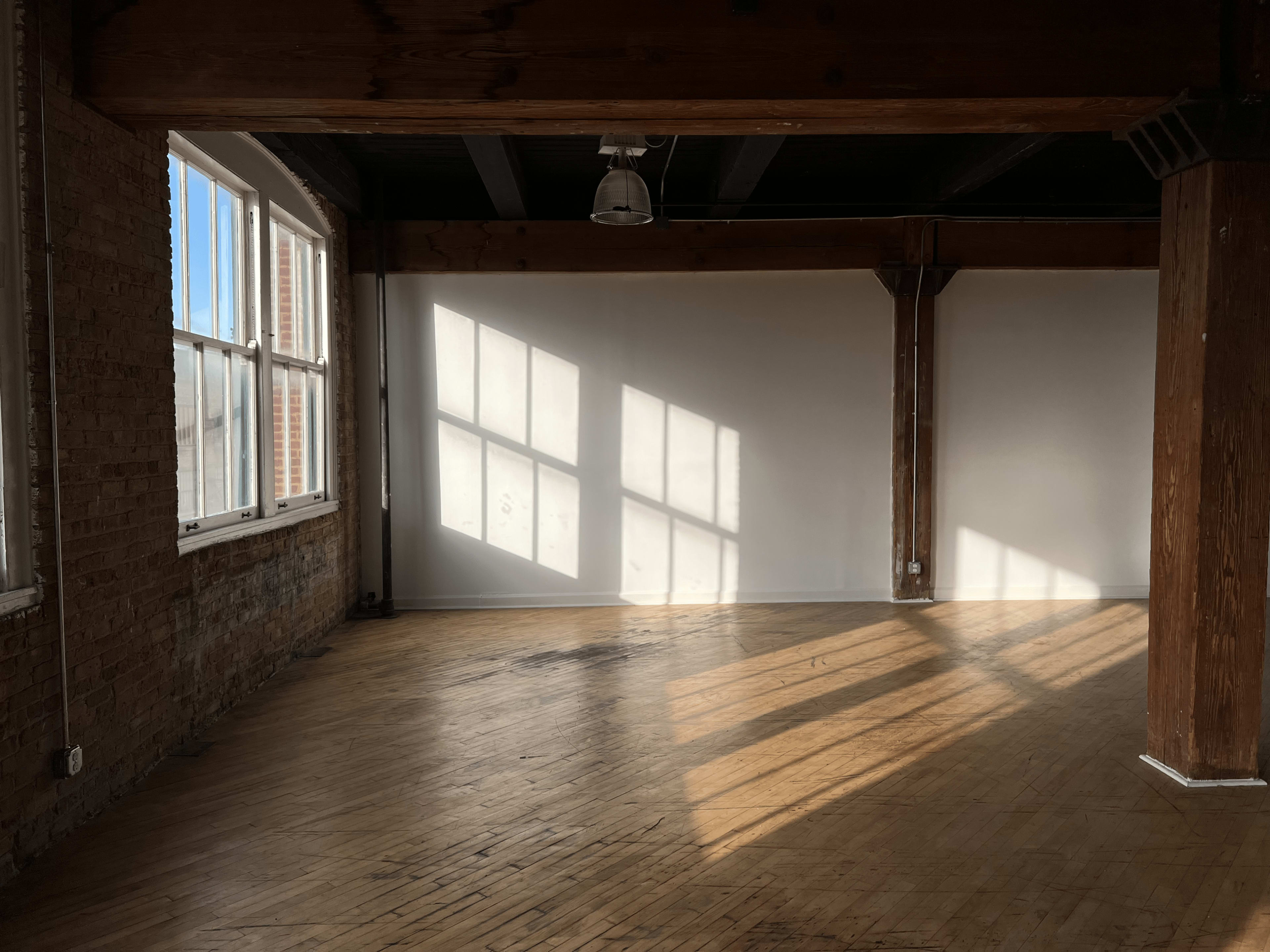 A spacious room with wooden floors and large windows casting shadows on the white walls.