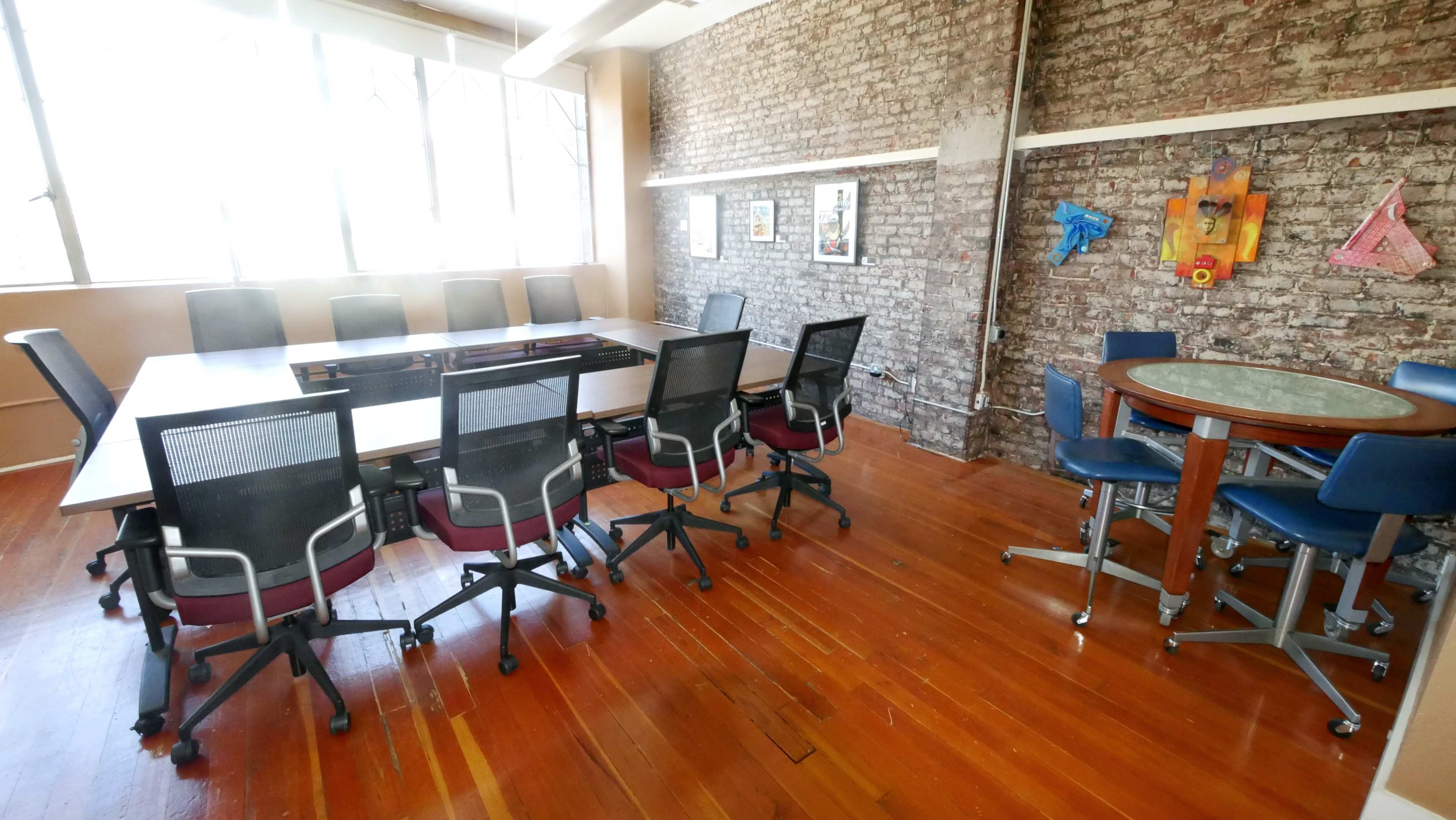 A conference room with a long table surrounded by chairs, large windows, and a brick wall.