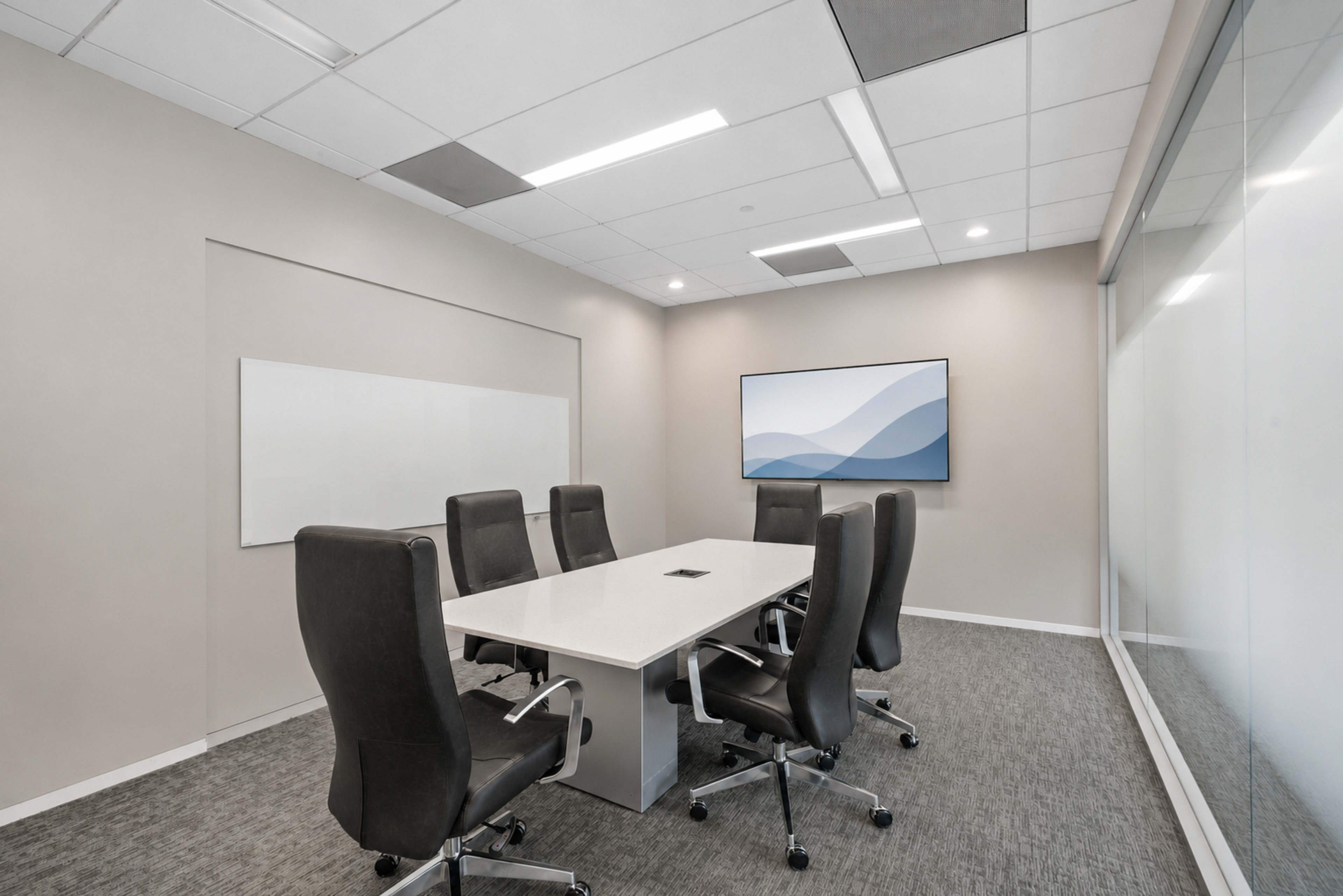 A modern conference room features a long white table surrounded by black office chairs, with a large screen mounted on the wall and glass panels on one side.