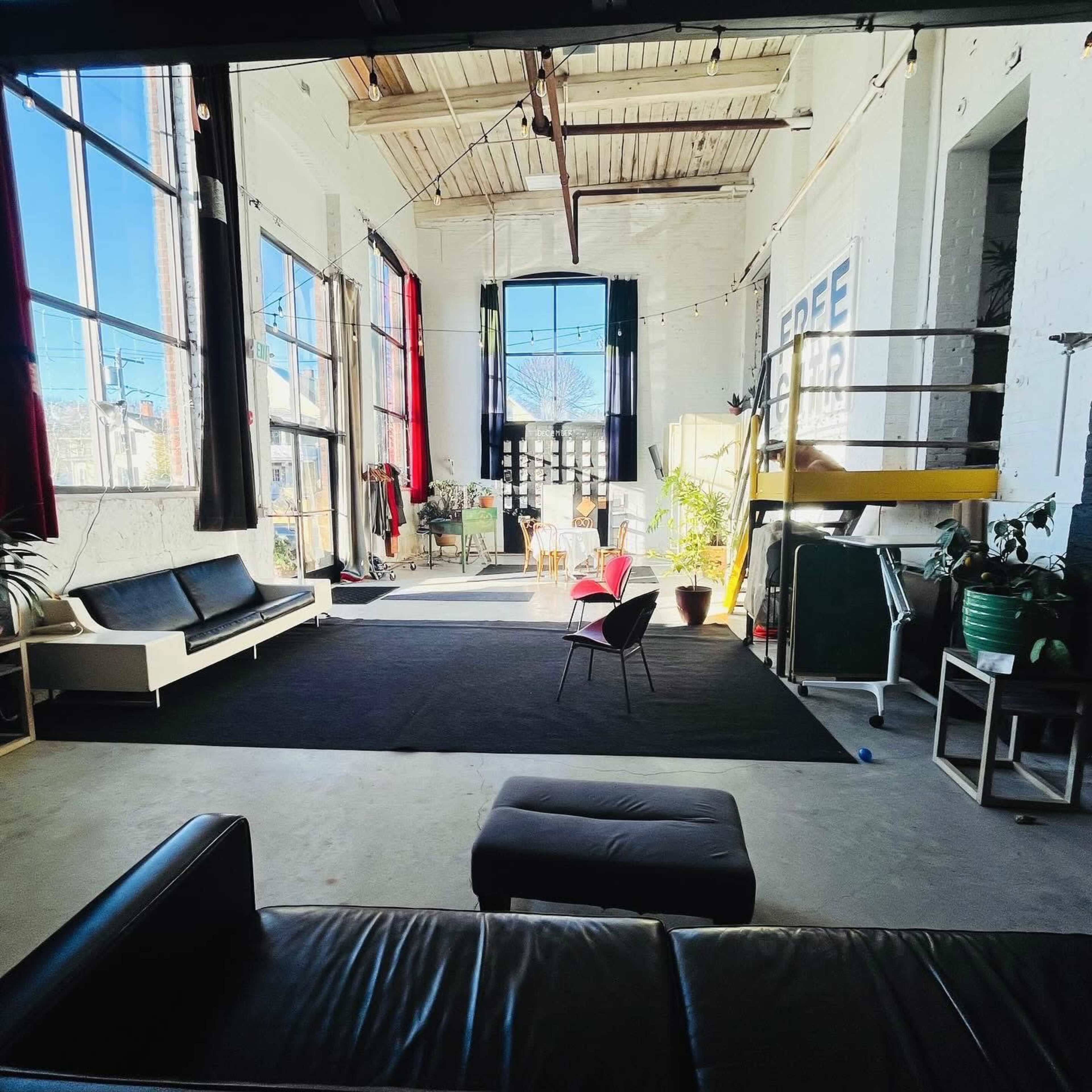 The image shows a spacious, industrial-style room with large windows, a black rug, seating areas, and a staircase leading to a loft.