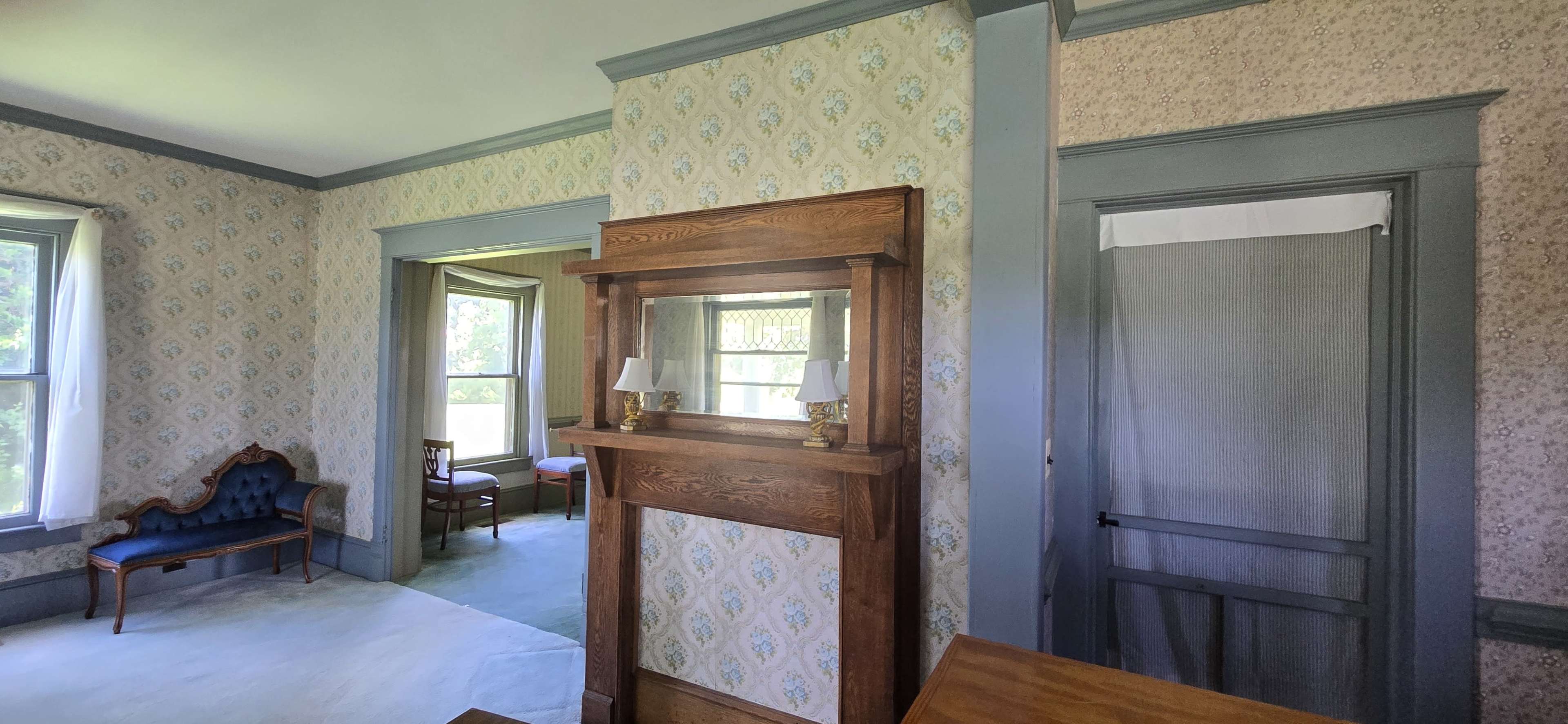 The image shows a vintage room featuring floral wallpaper, an ornate wooden fireplace with lamps, and various pieces of period furniture including a blue upholstered chair.