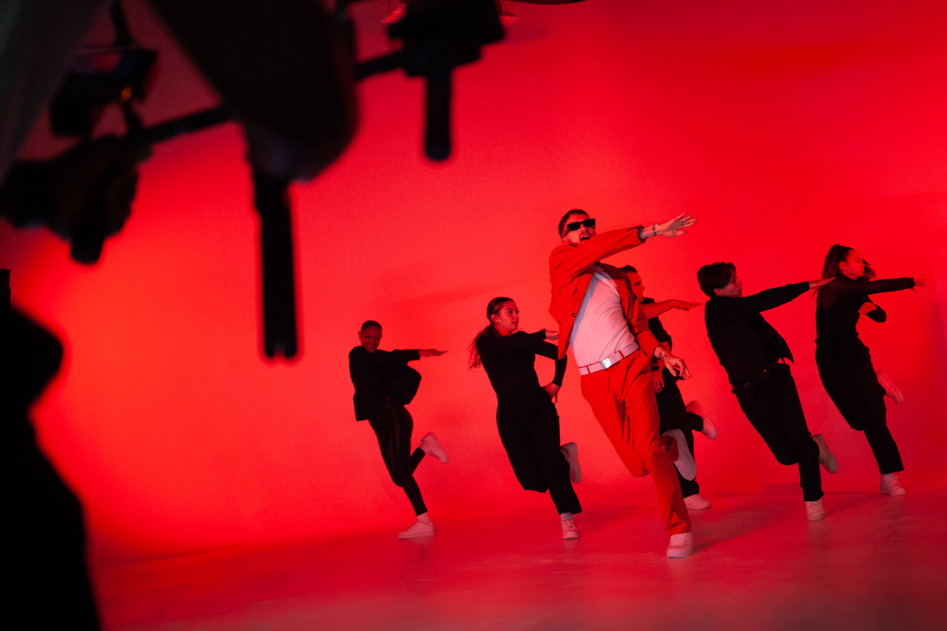 A group of dancers performs in unison against a bright red backdrop, with one dancer in a red outfit leading the formation.