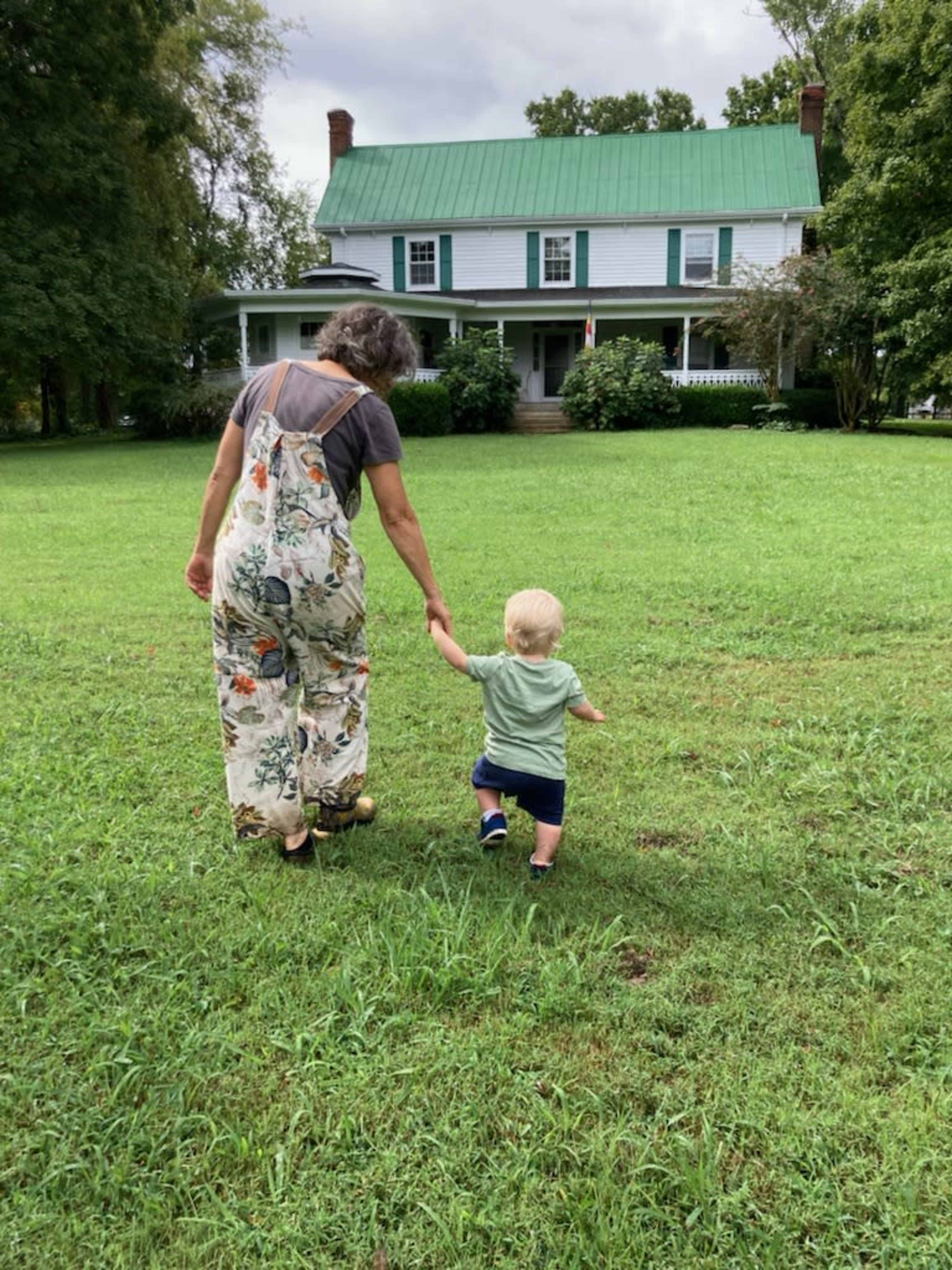 A person and a young child walk hand-in-hand across a grassy area towards a large house with a green metal roof.