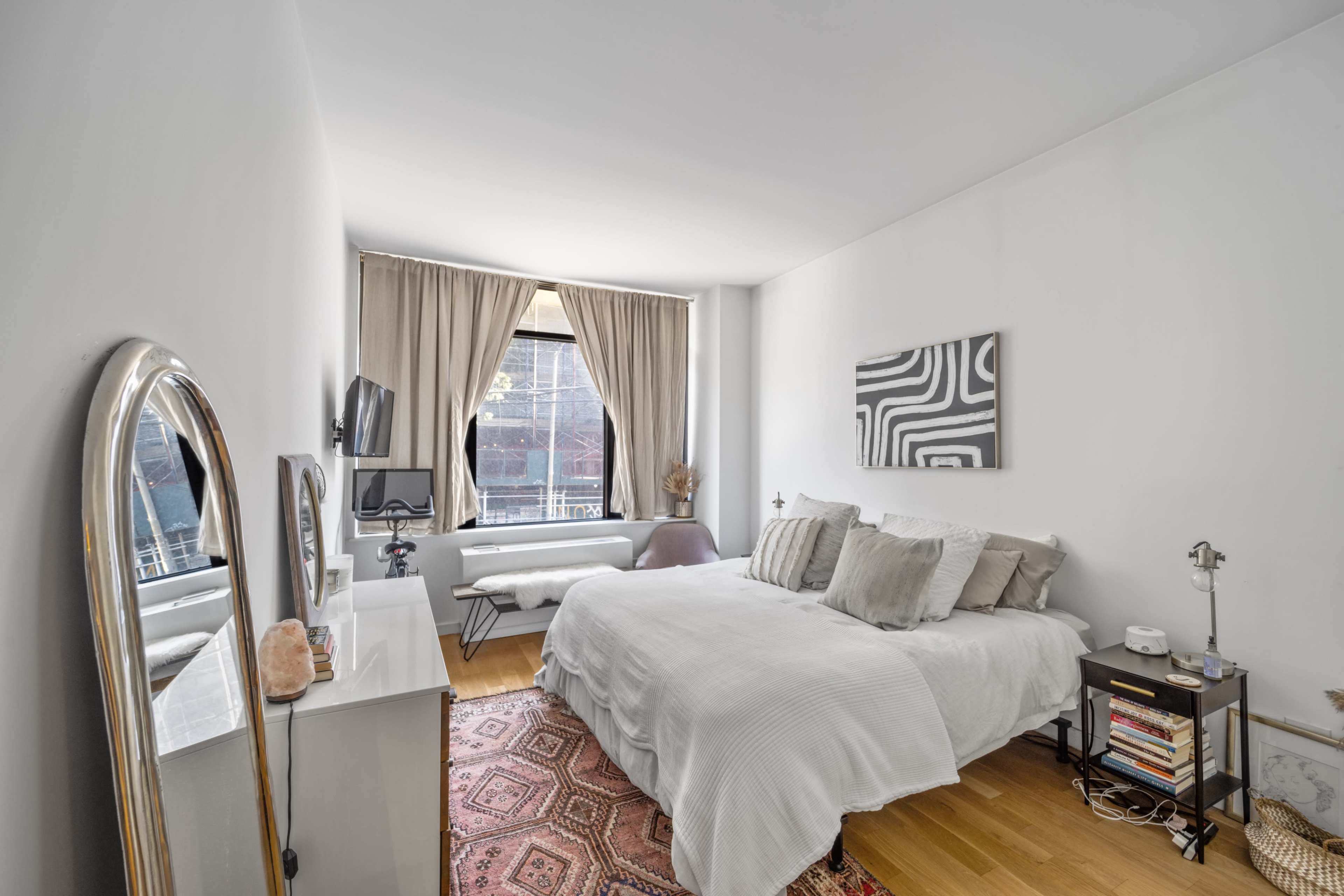 The image shows a modern bedroom with a white bed, a large mirror, a television mounted on the wall, and sheer curtains covering a window.
