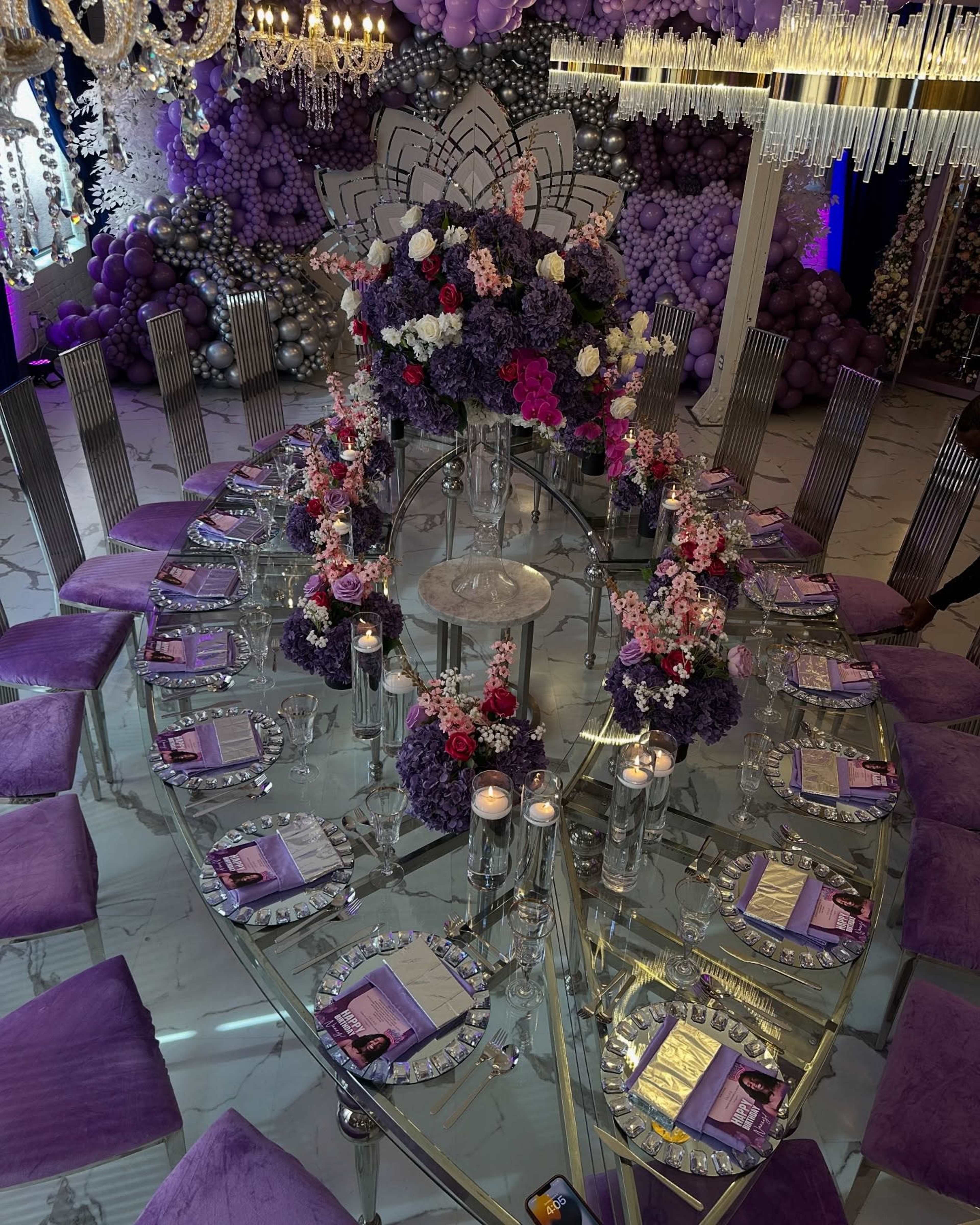 A large, elegantly set dining table with purple accents features circular floral centerpieces and a backdrop of balloon decorations.