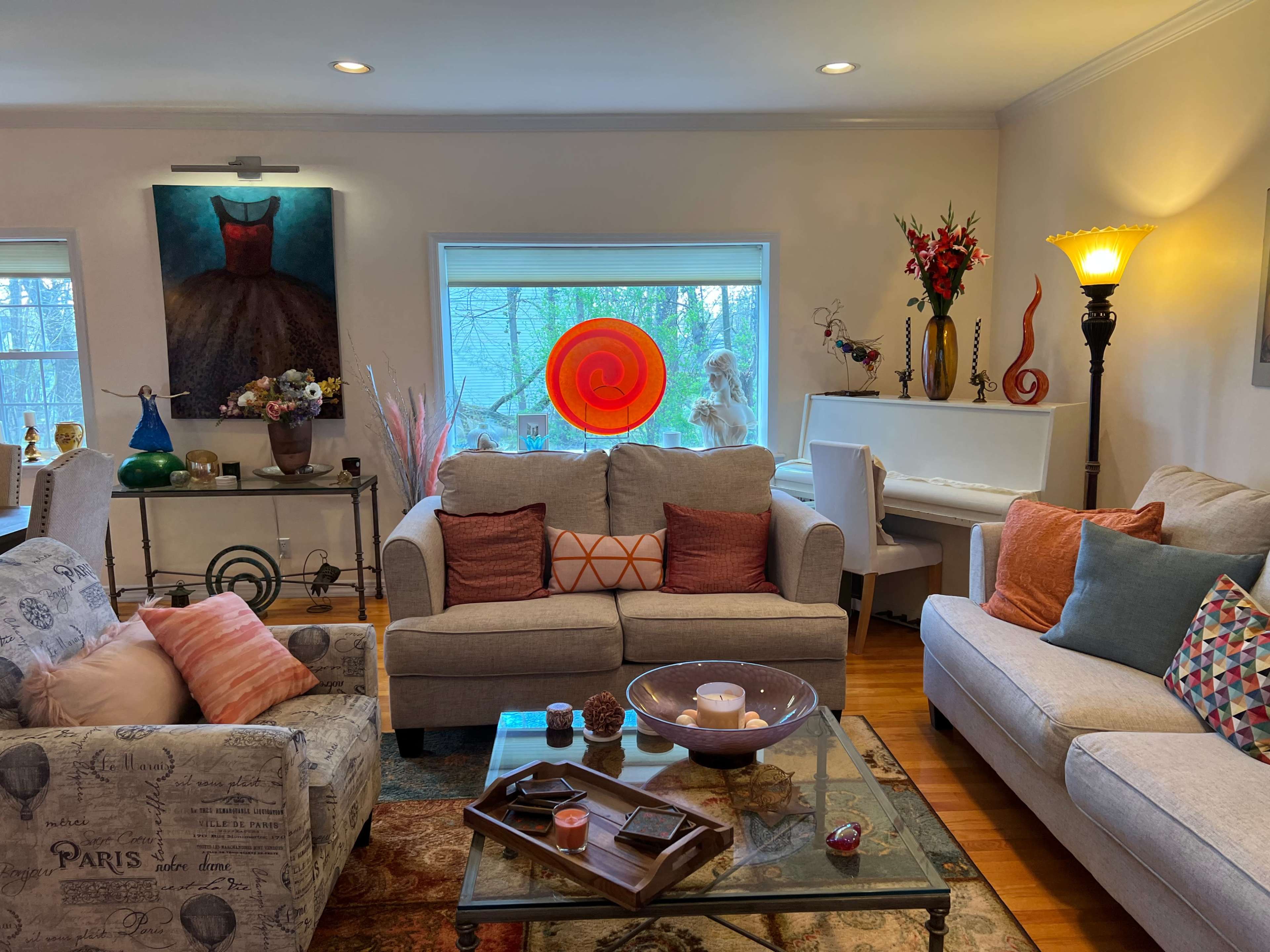 The living room features two sofas, a glass coffee table, a piano, and large windows showing a view of trees outside.