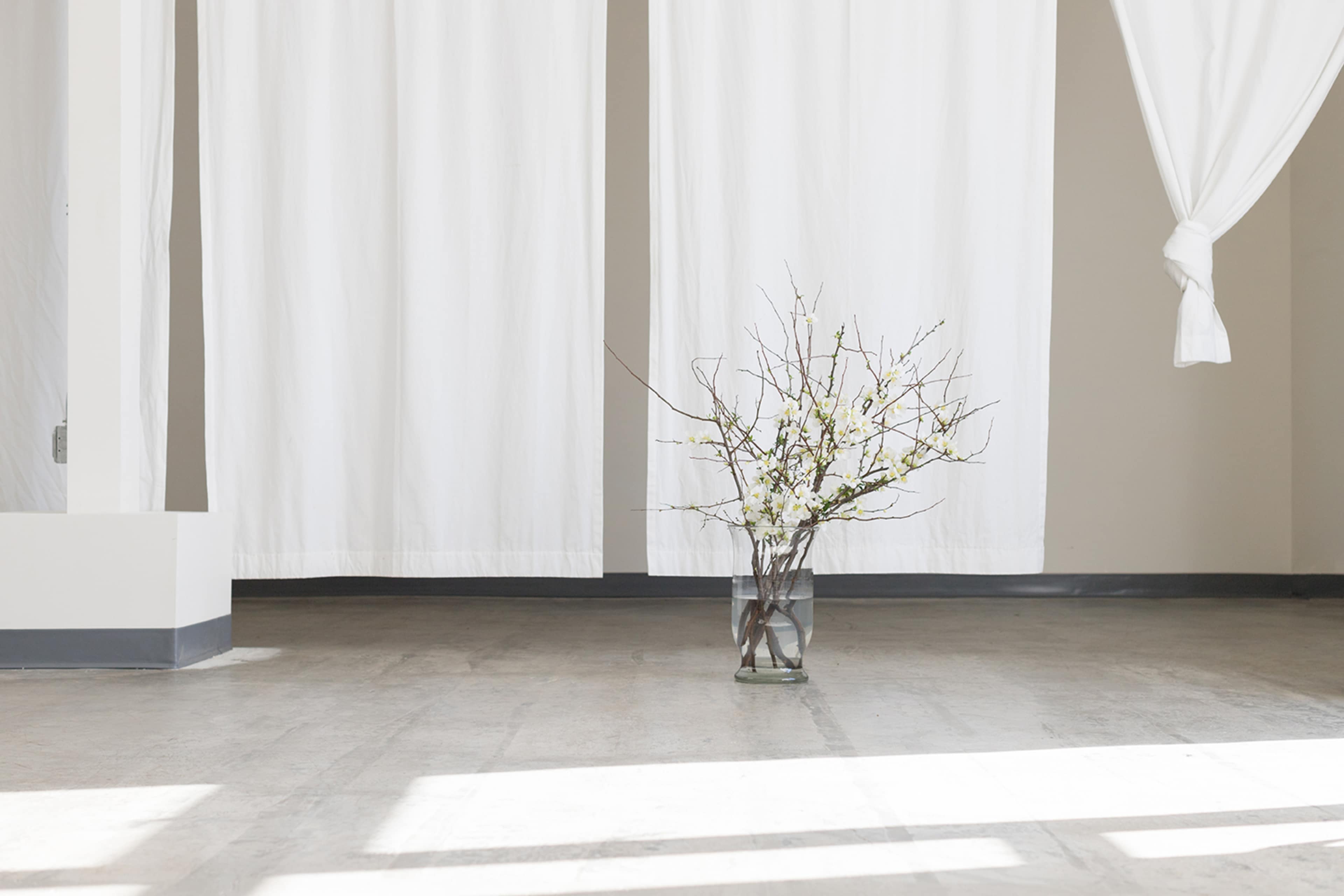 A glass vase with branches and flowers is placed in the center of a room with white draped curtains.
