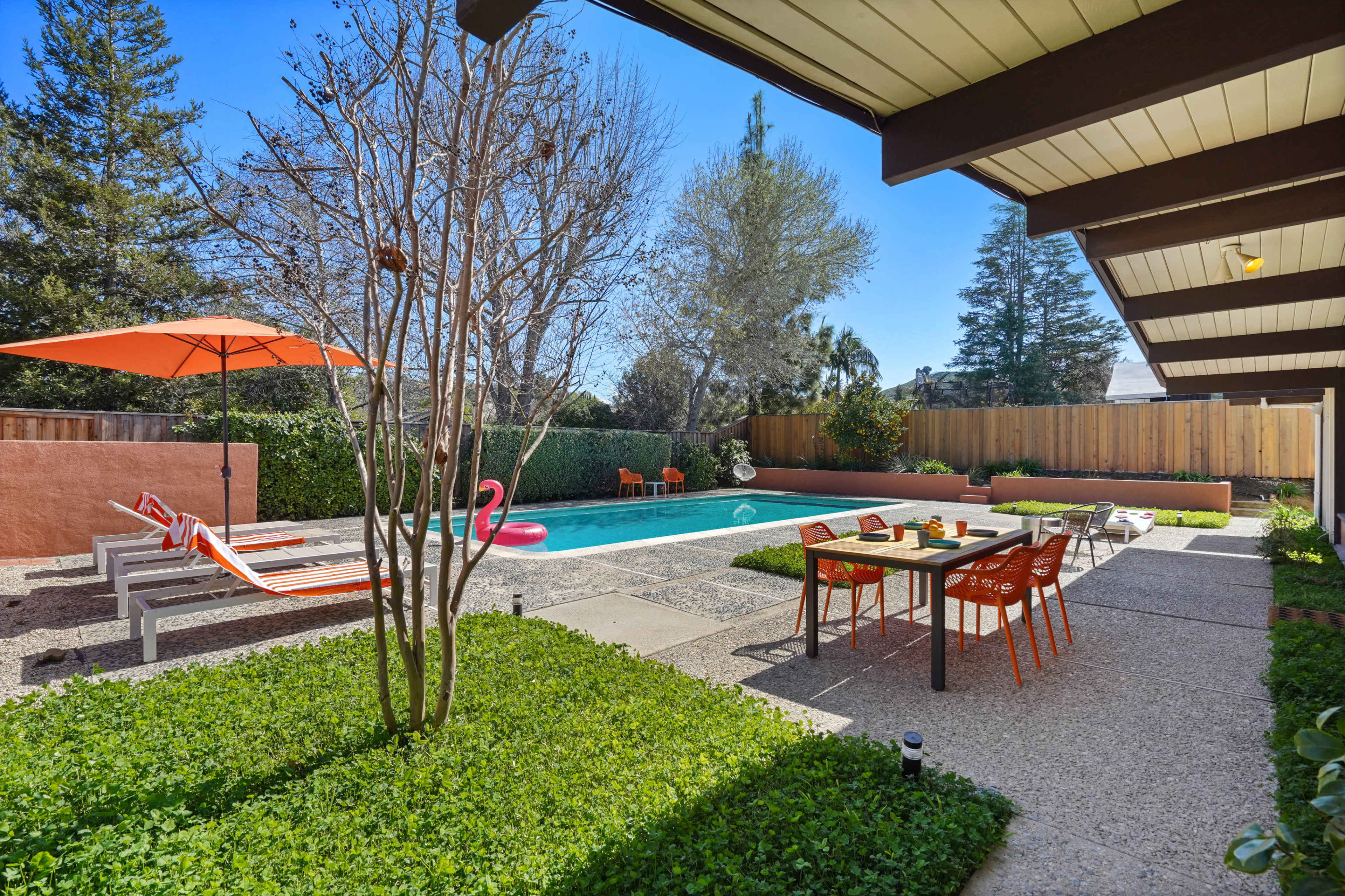 The image shows a backyard with a swimming pool, lounge chairs, an orange umbrella, and a dining table surrounded by greenery.