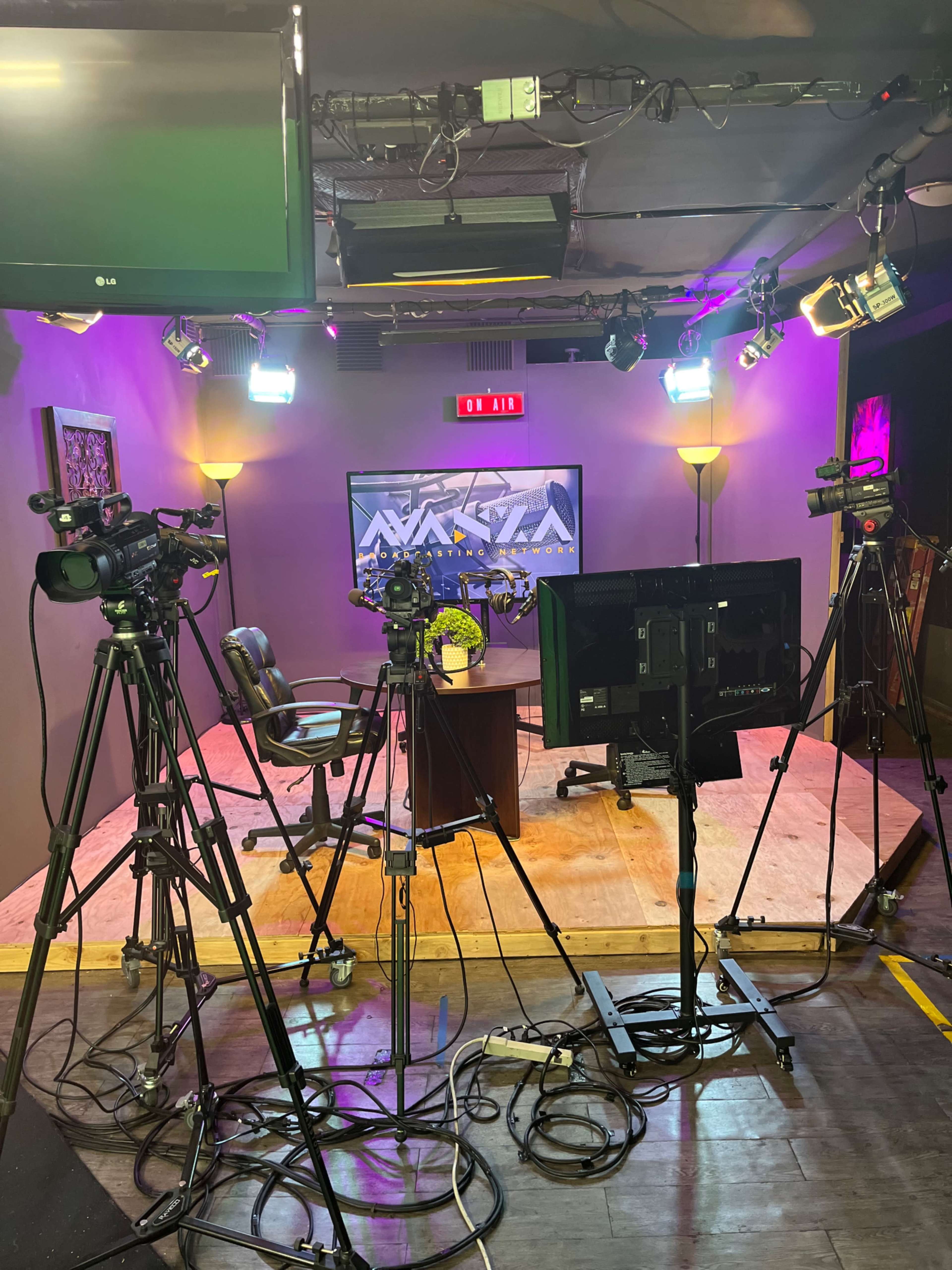 A television studio setup with multiple cameras positioned around a central desk and display screen, illuminated by various studio lights.