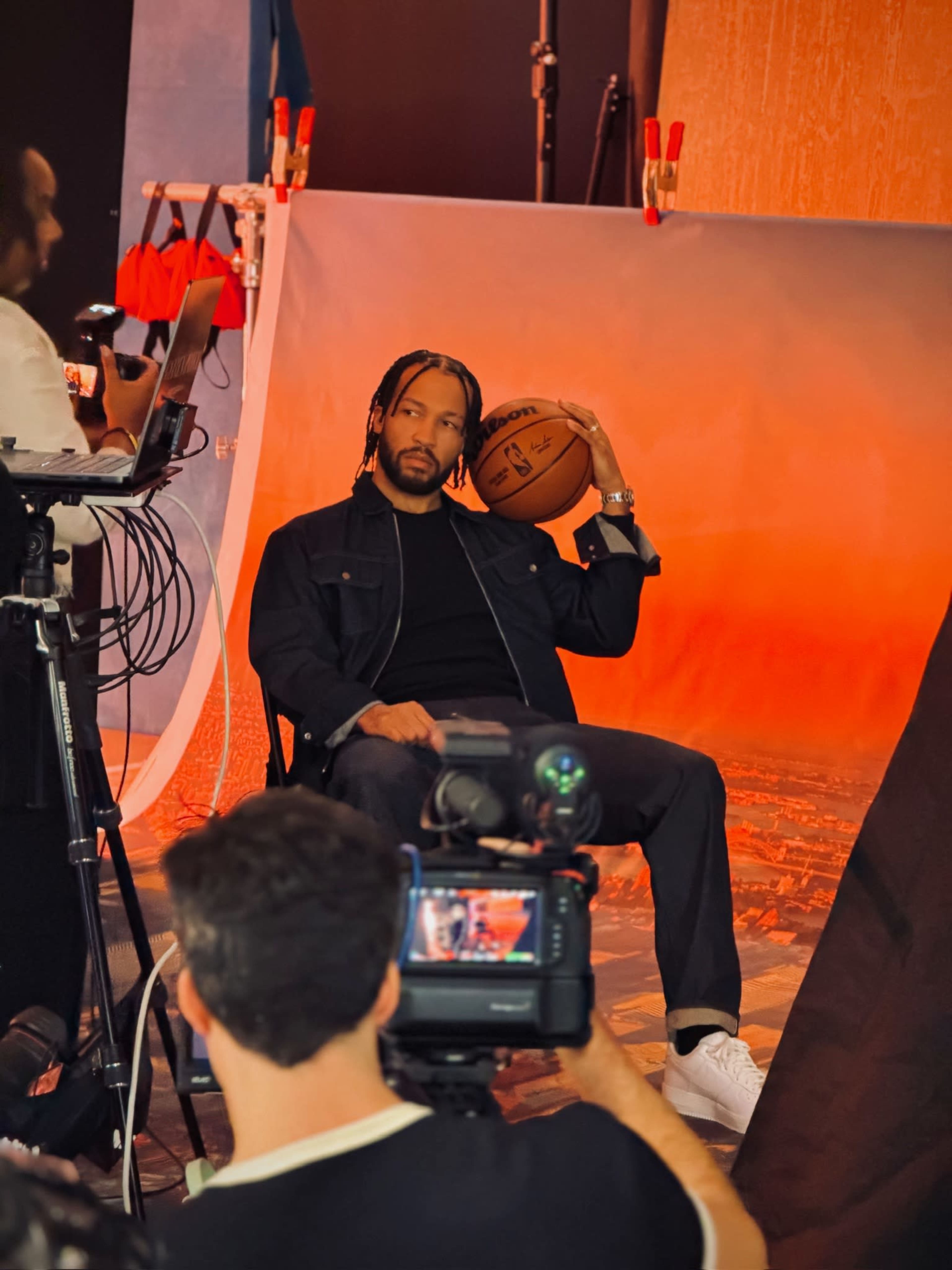 A person sits on a chair holding a basketball in a photoshoot setting with a colorful backdrop and cameras present.