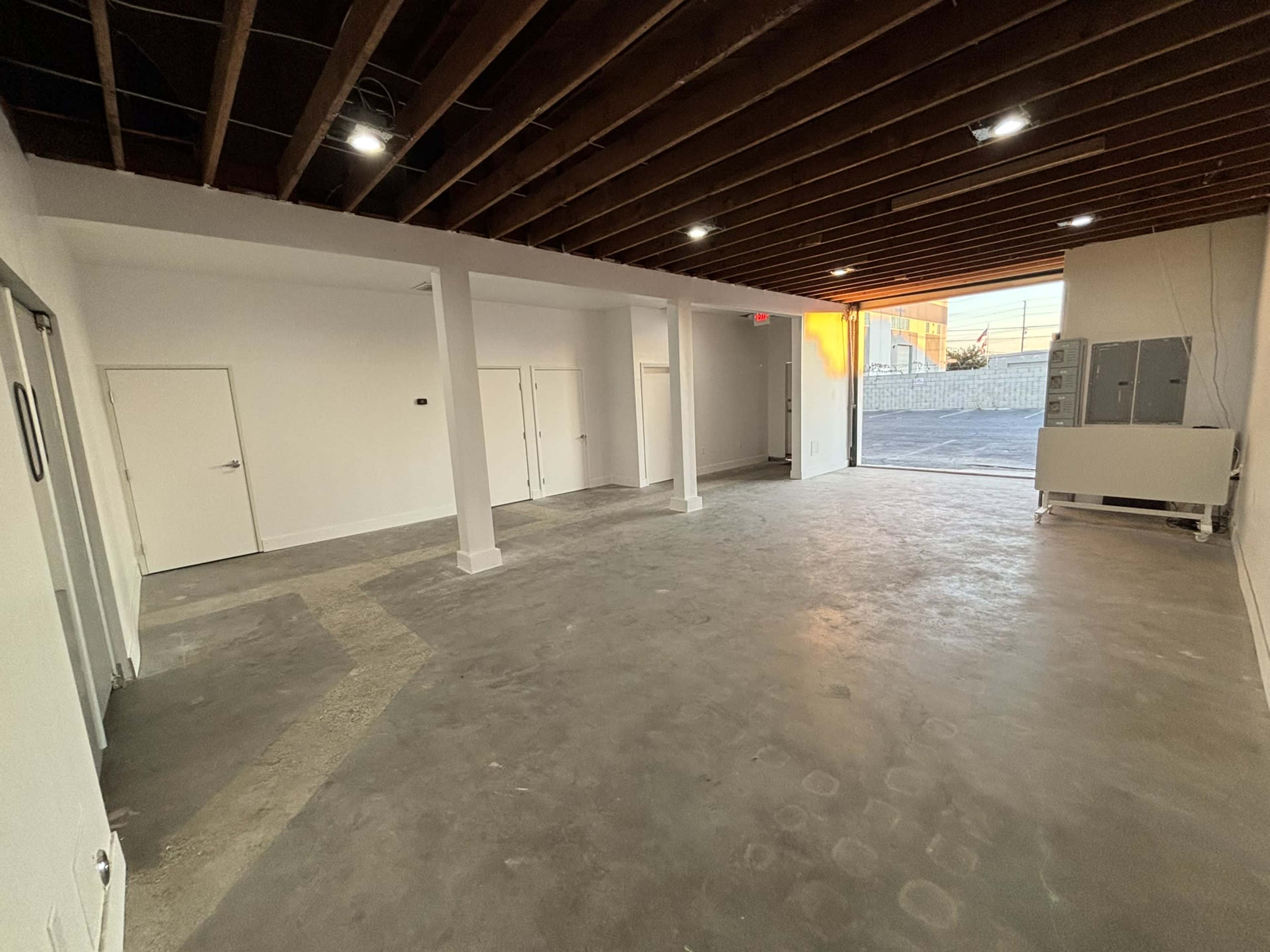 The image shows a spacious, empty room with concrete flooring, white walls, and exposed wooden beams on the ceiling, featuring multiple doors and an entrance illuminated by natural light.