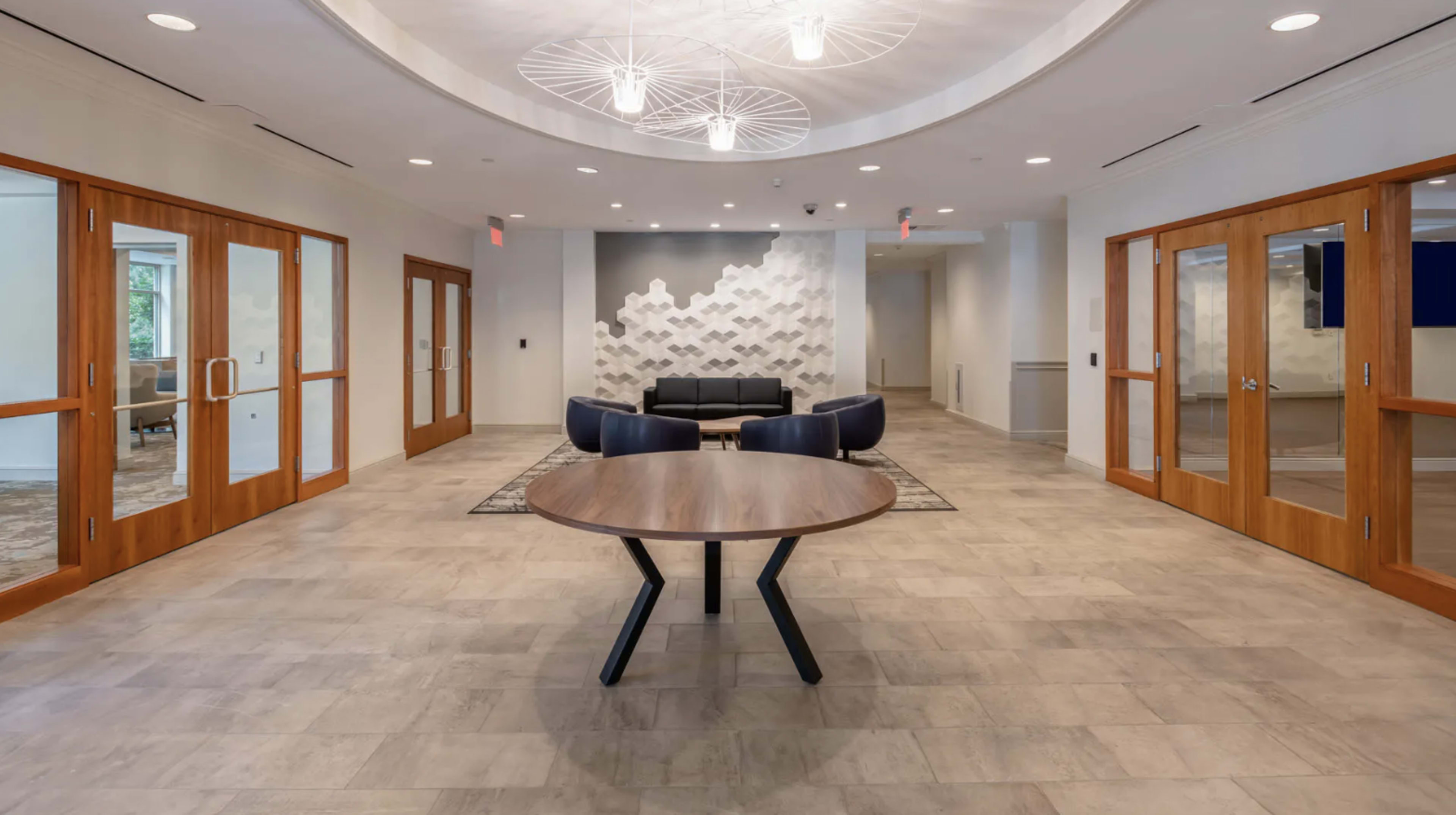 The image shows a modern lobby area with a round table in the center, surrounded by two blue chairs and a black couch, with decorative wall features and large windows.