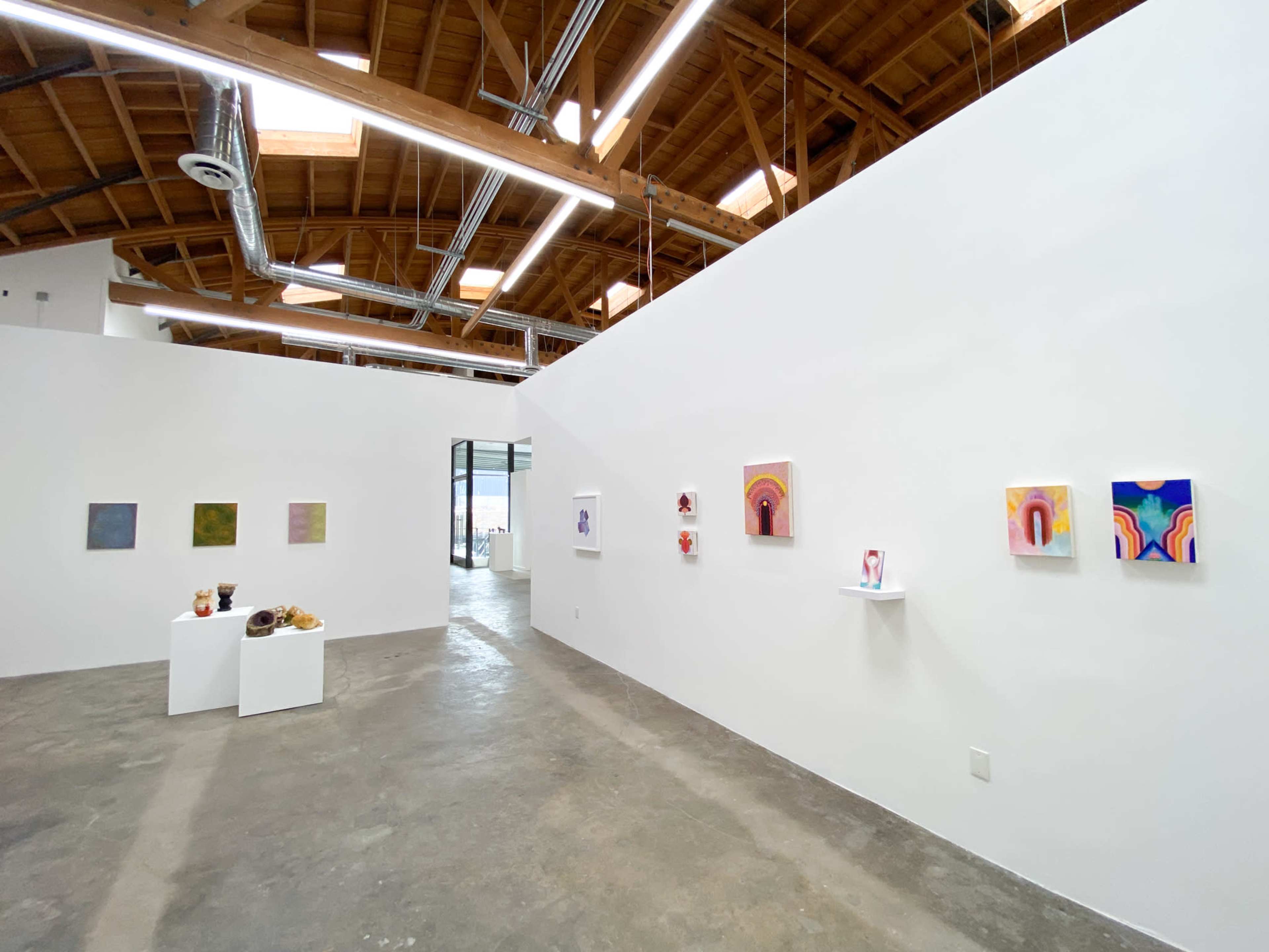 An art gallery with white walls featuring various paintings and small sculptures displayed on shelves and a table.