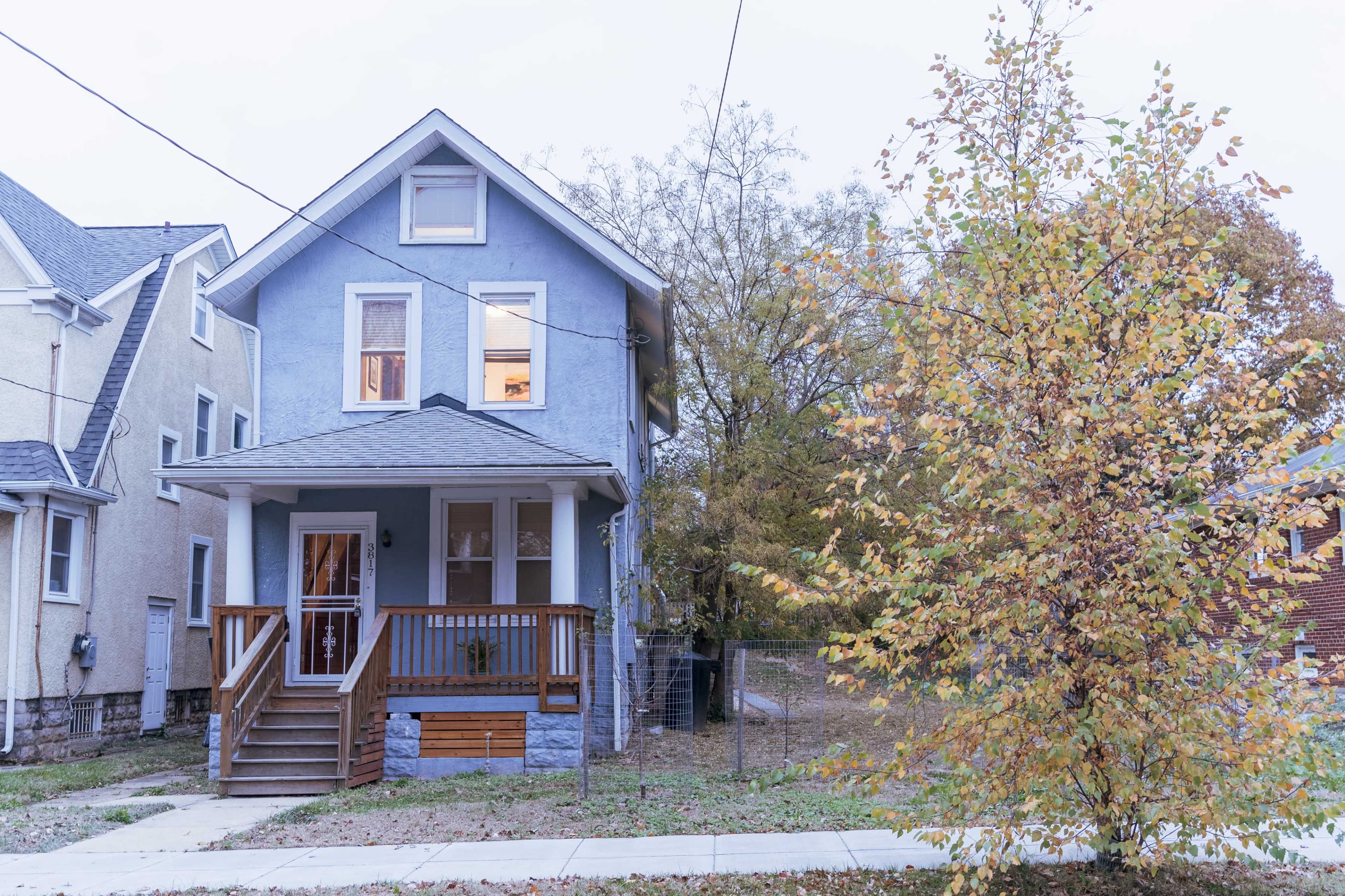 A two-story blue house with a wooden front porch is set beside a small tree with yellow leaves in a residential area.