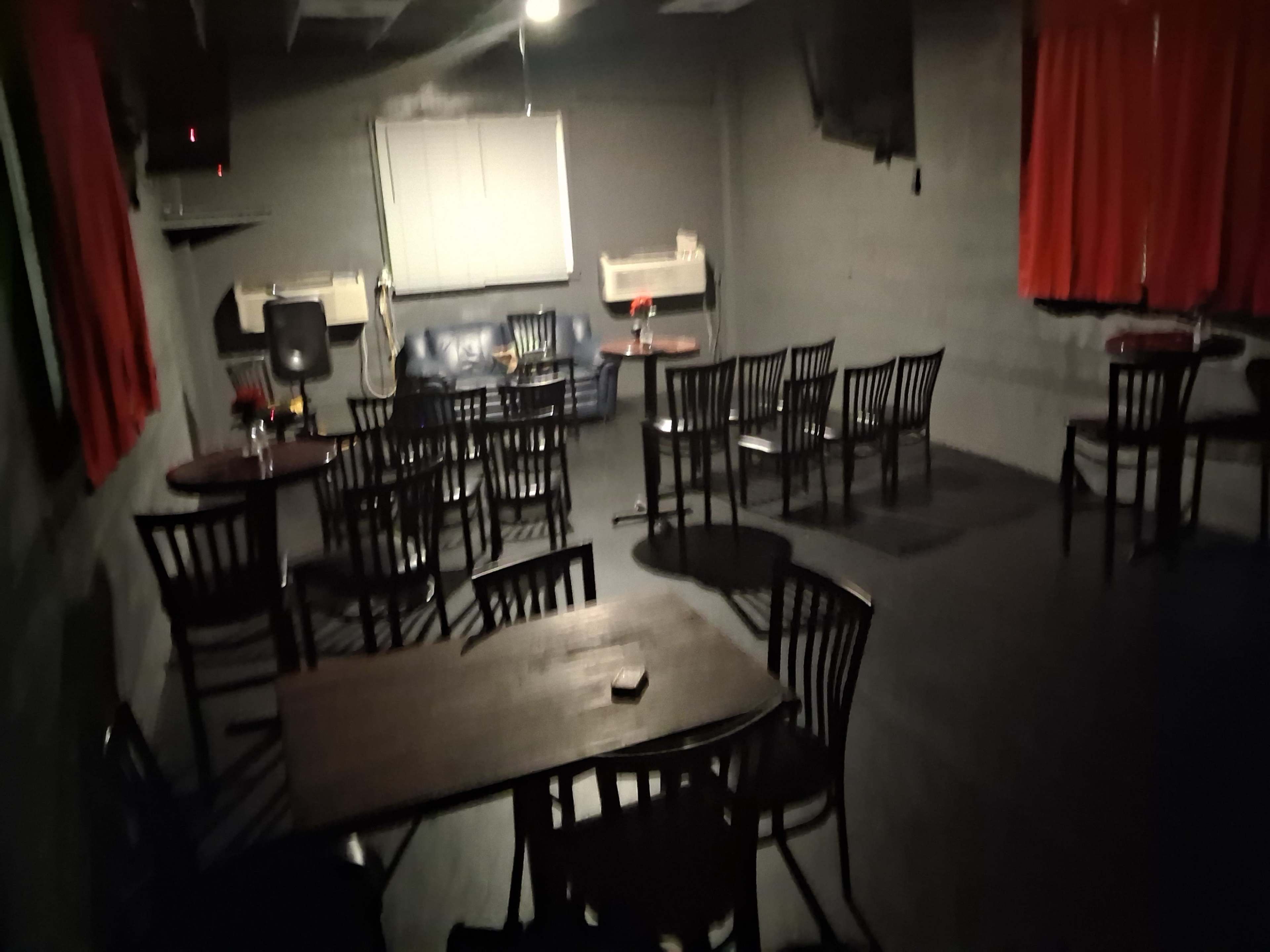 The image shows a dimly lit room set up with several chairs and tables, with red curtains on the walls and a few appliances in the background.