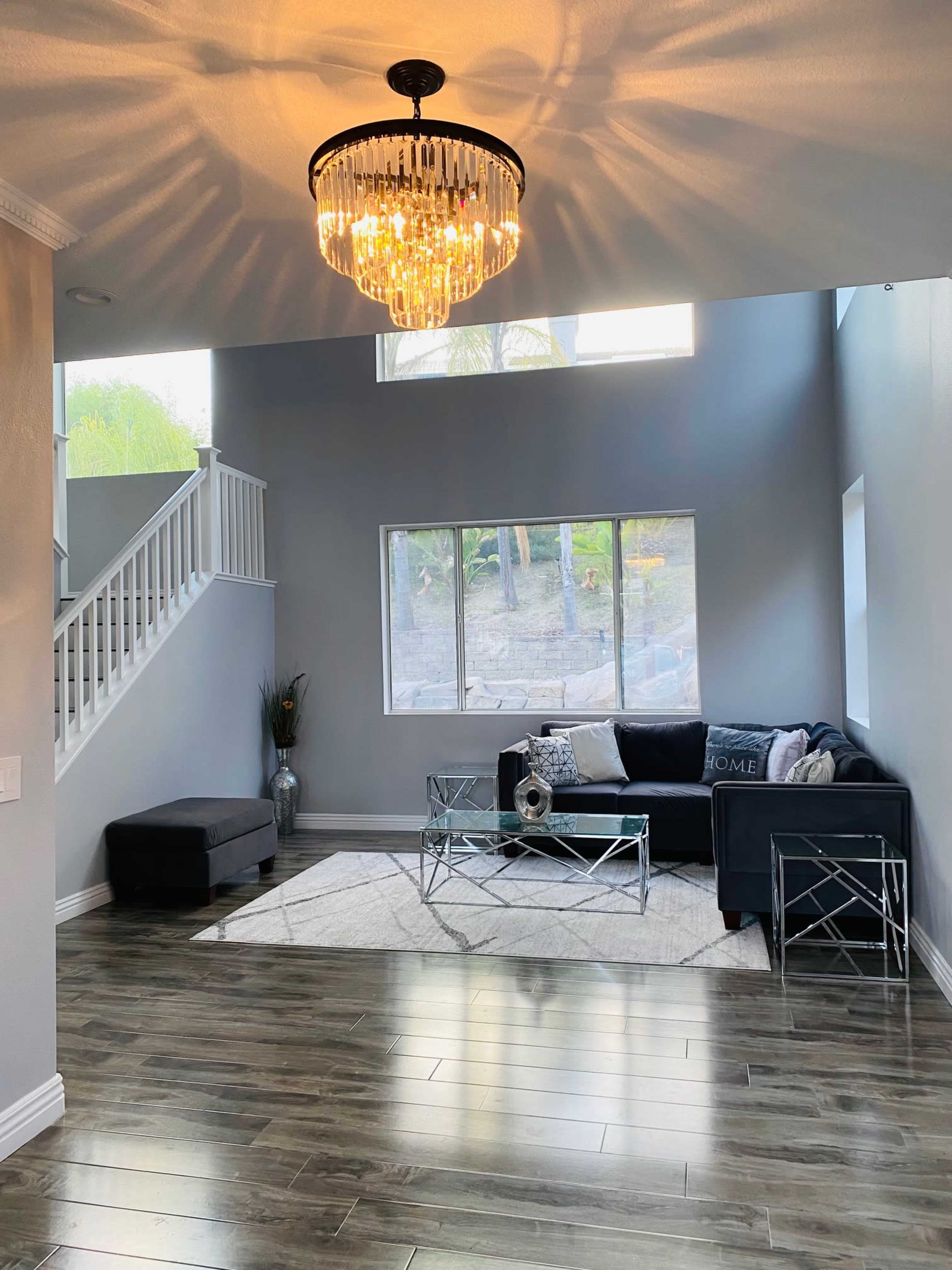 A modern living room features a chandelier illuminating a dark sectional sofa and glass coffee table, with large windows providing natural light.