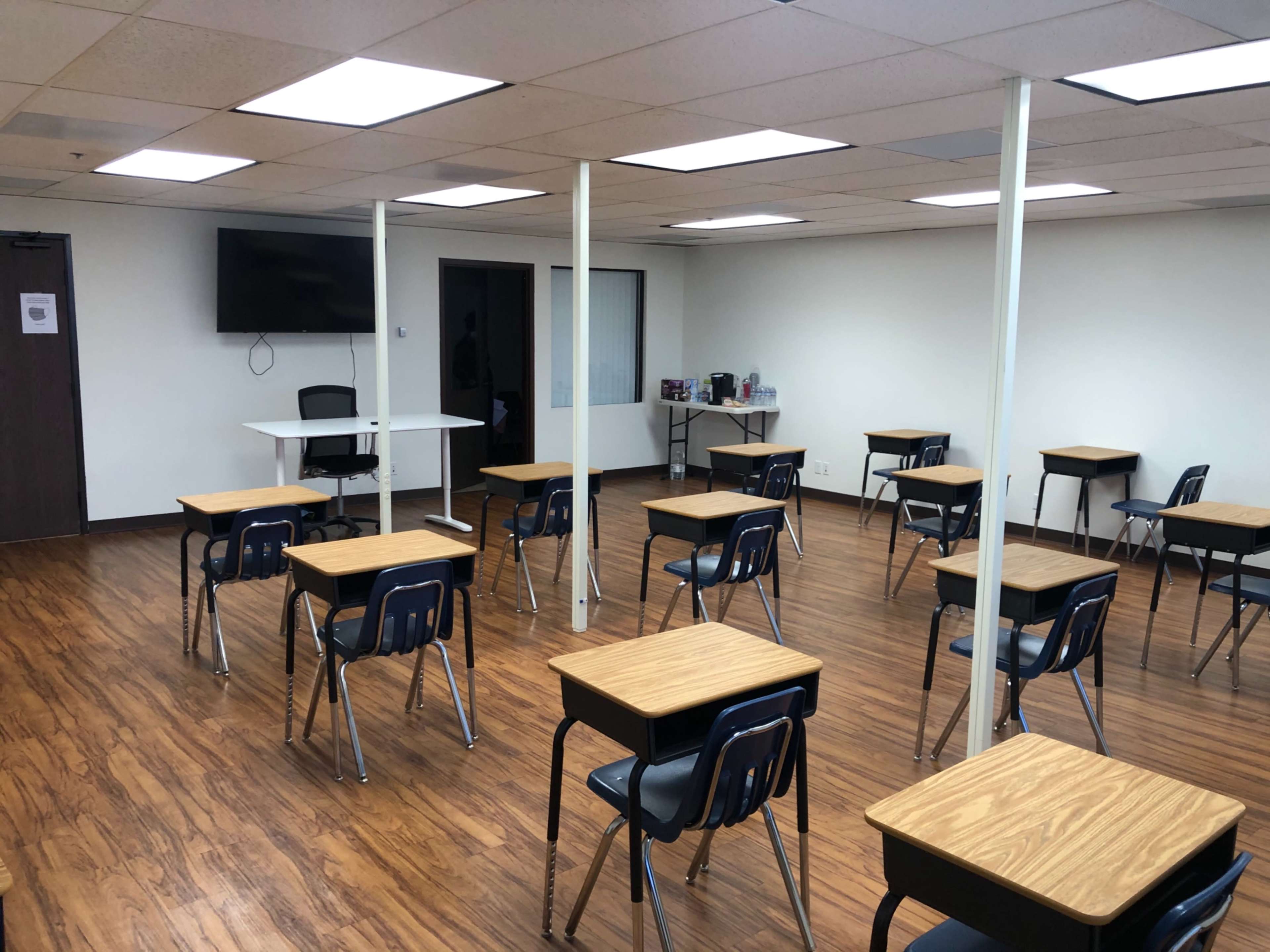 The image shows an empty classroom with several desks arranged in rows, a television on the wall, and a small table at the front.