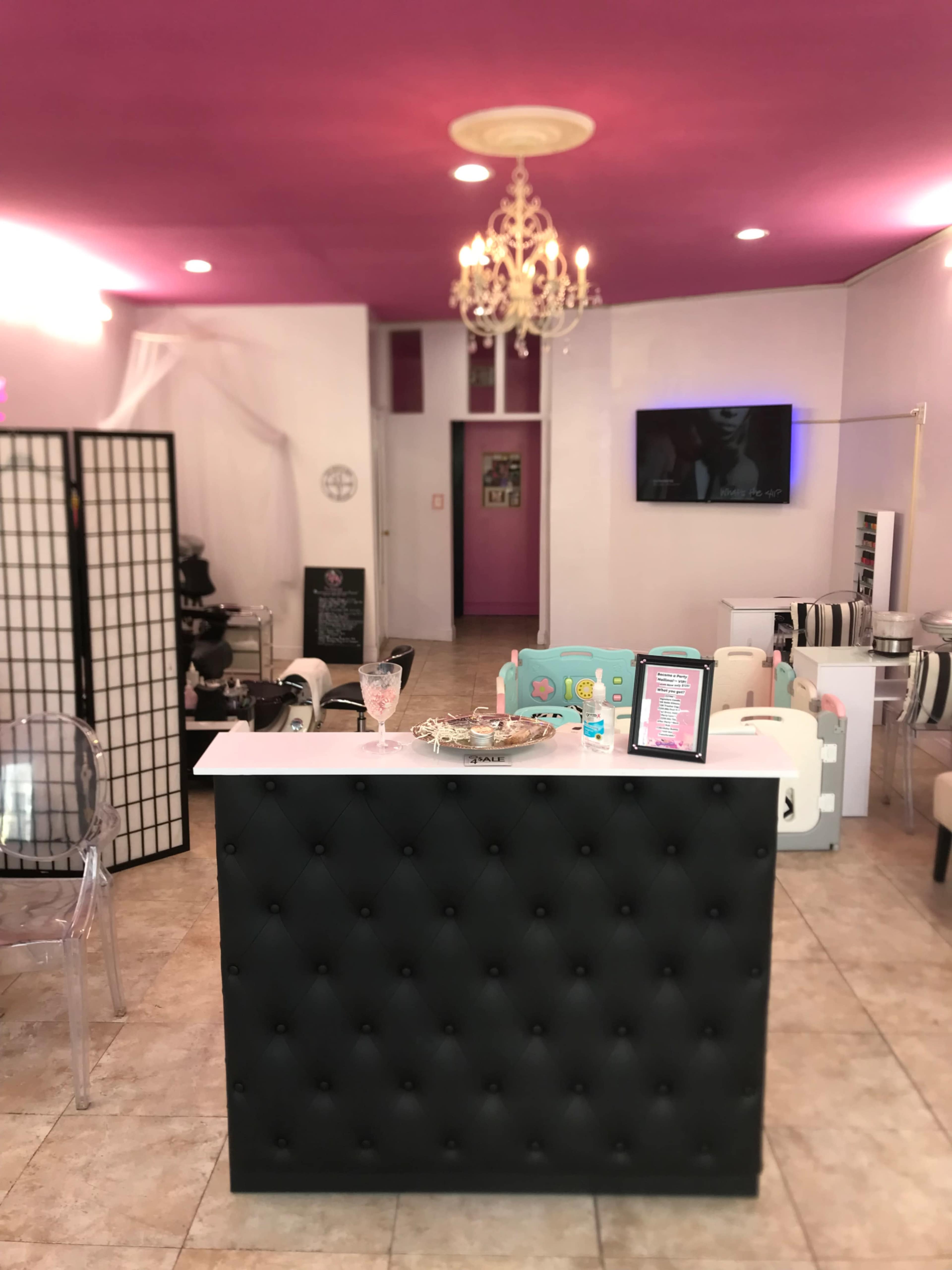 A beauty salon's reception area featuring a black tufted counter, pink walls, a chandelier, and various salon equipment.