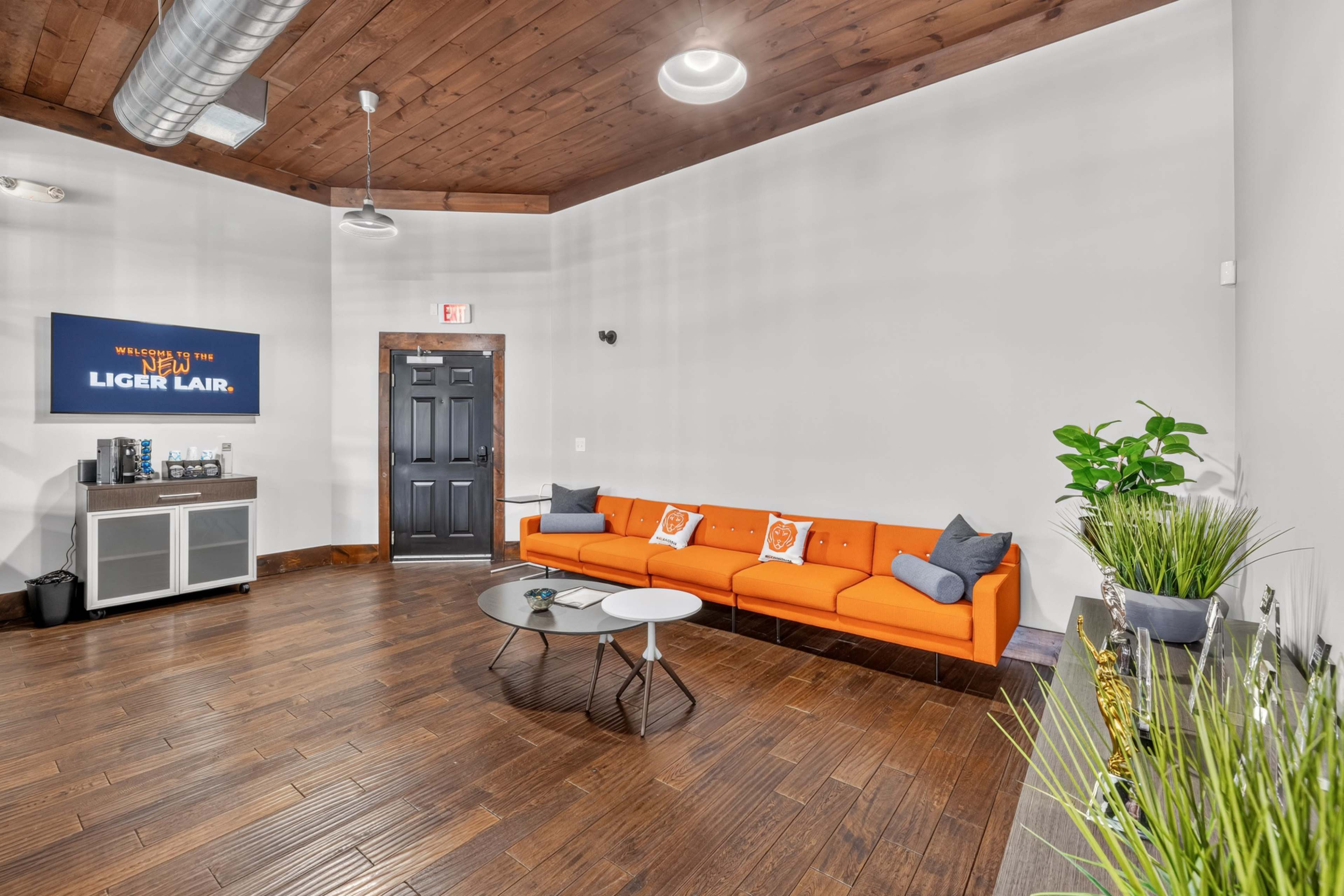 The image shows a modern lounge area with an orange sectional sofa, a circular coffee table, and a small plant, all set against a wooden ceiling and floor.
