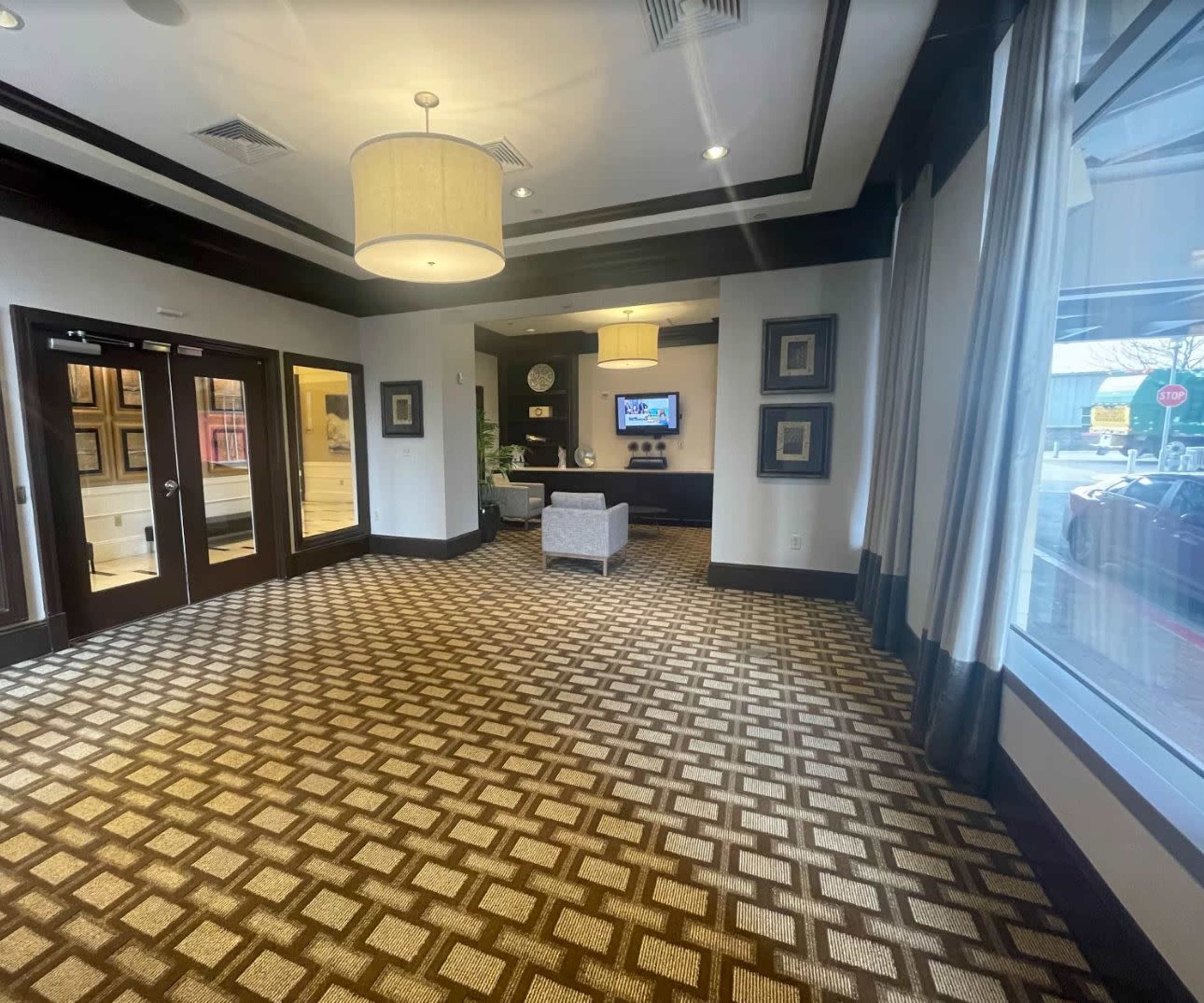 A spacious hotel lobby with patterned carpet, a seating area, and large windows letting in natural light.