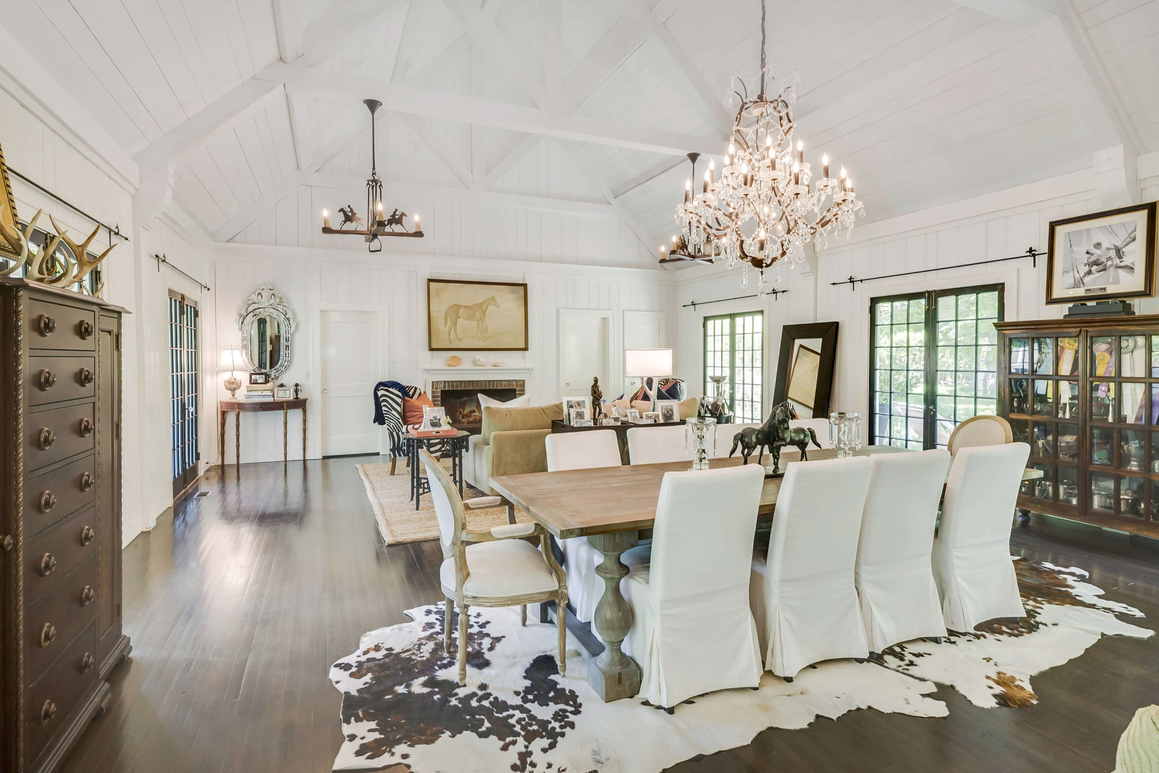 The image depicts a spacious dining room featuring a large wooden table surrounded by white upholstered chairs, with a chandelier overhead and a mix of decorative elements and artwork on display.