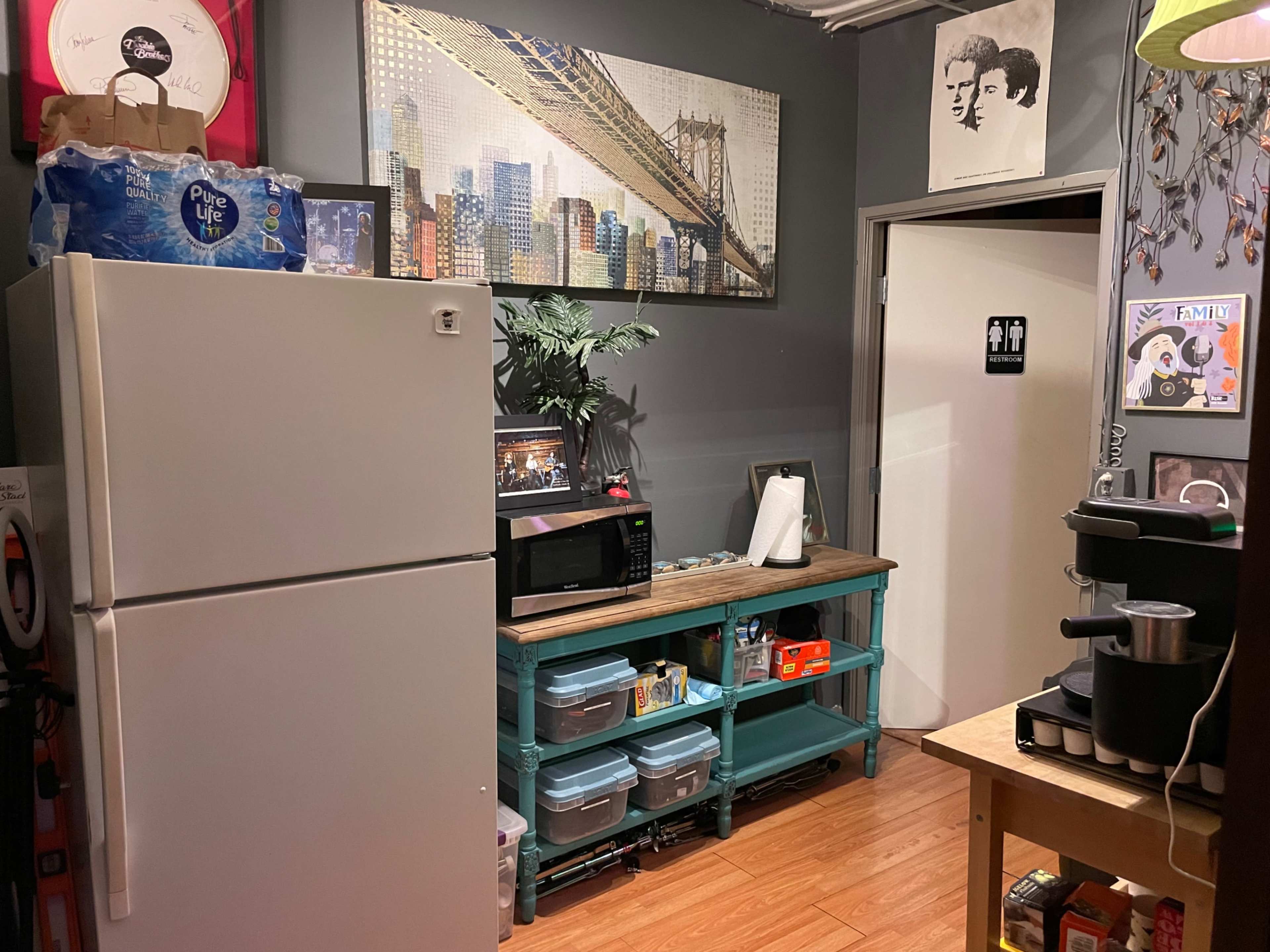 The image shows a small kitchen area featuring a refrigerator, a microwave, a wooden table with a coffee maker, and storage containers on a turquoise cart, with wall art and a plant in the background.