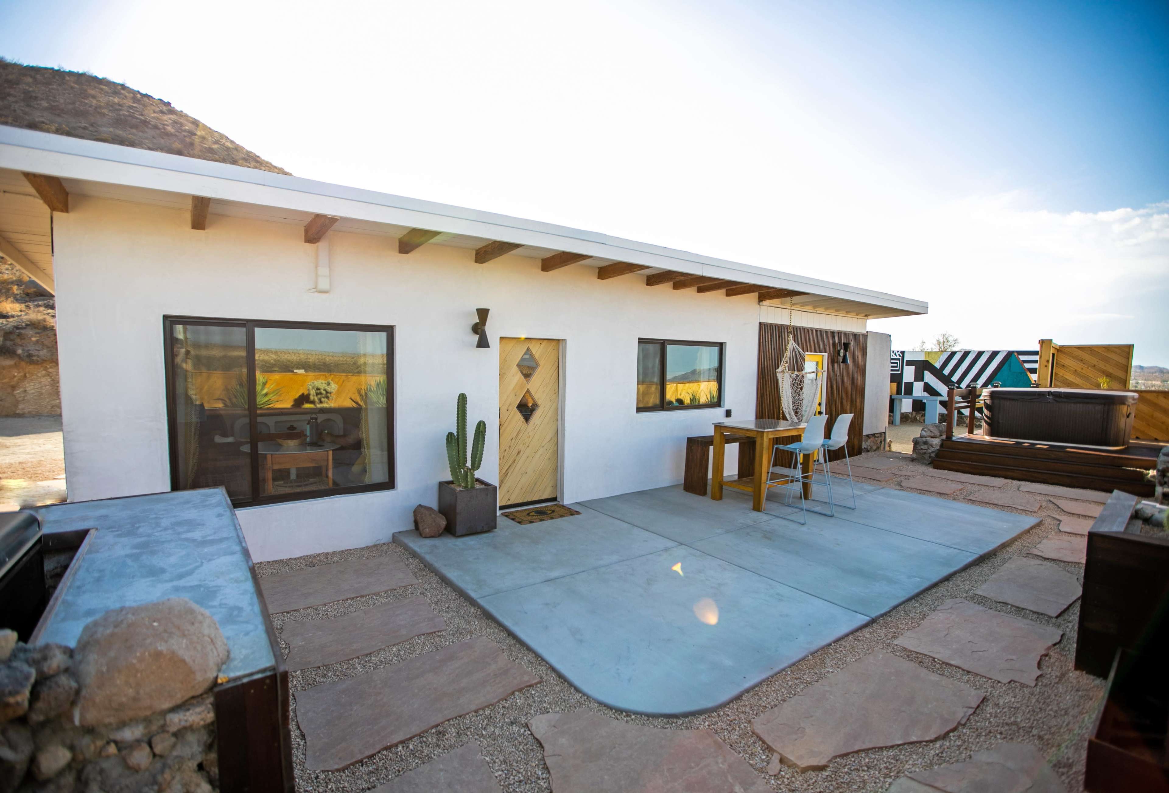 A modern desert house features a concrete patio, a wooden door, and outdoor seating surrounded by sparse landscaping.