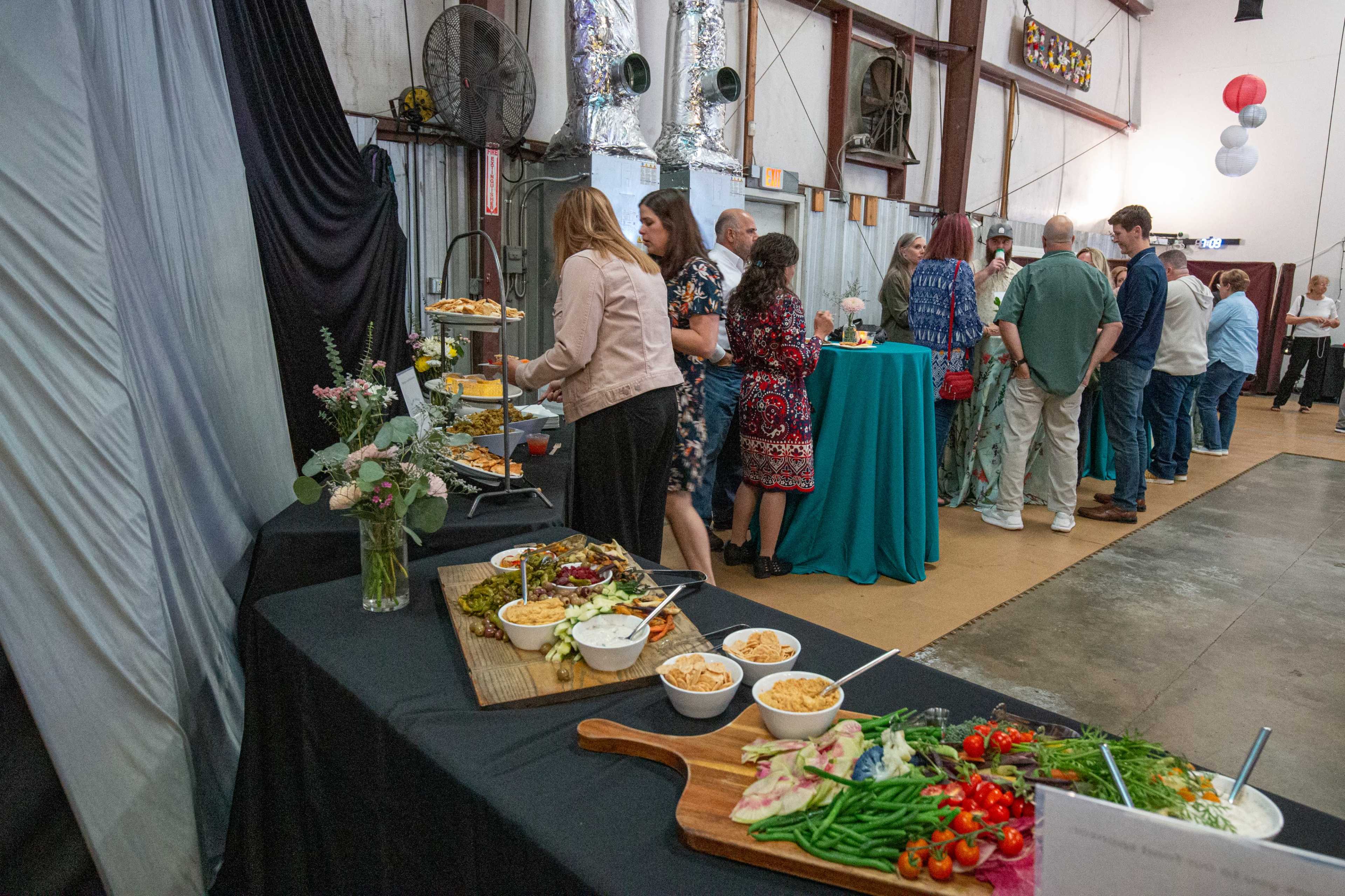 A group of people stands in line at a buffet table featuring a variety of foods, while others serve themselves from the spread.