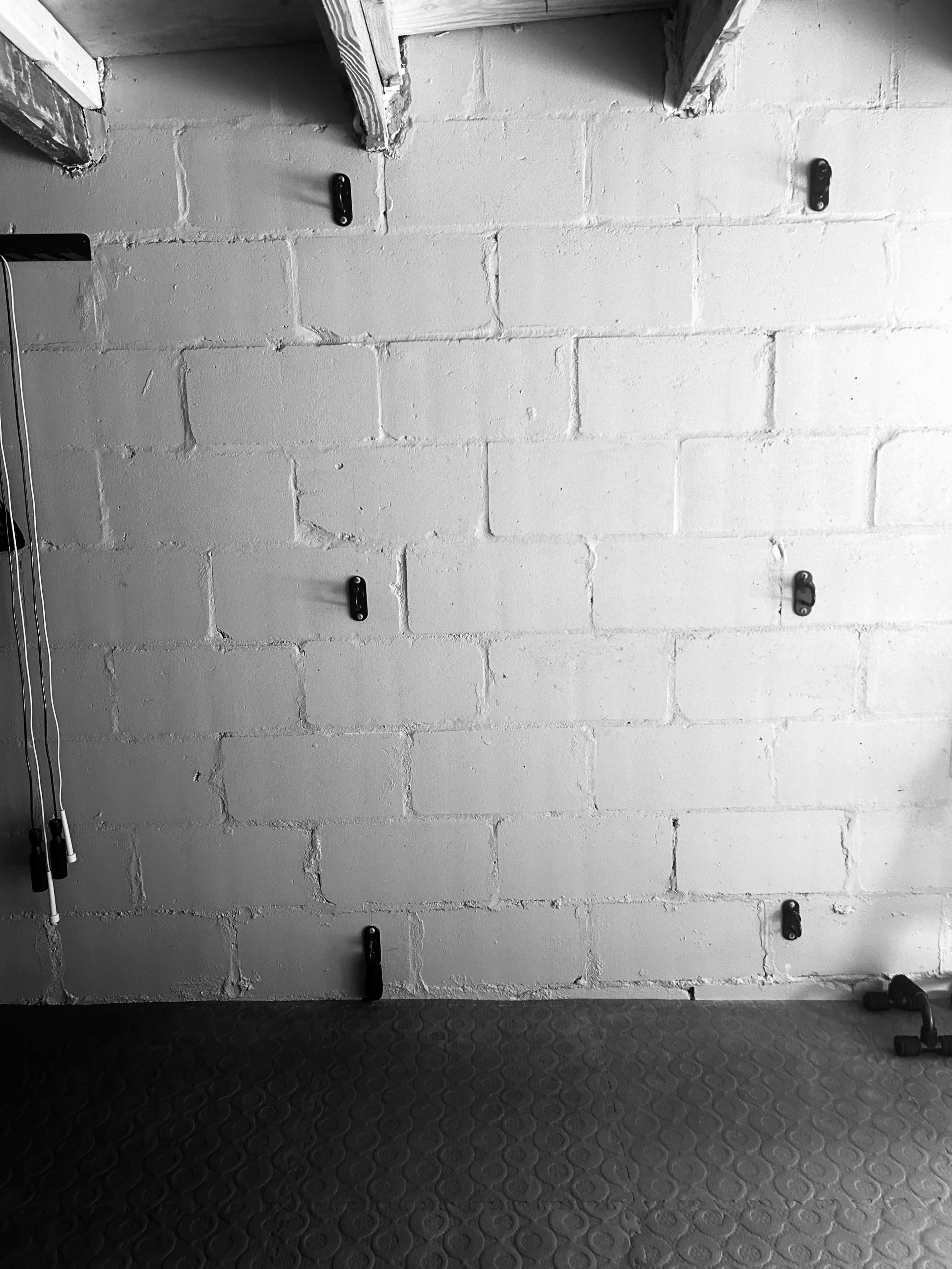 A bare concrete wall with several black hooks mounted on it and a textured floor below.