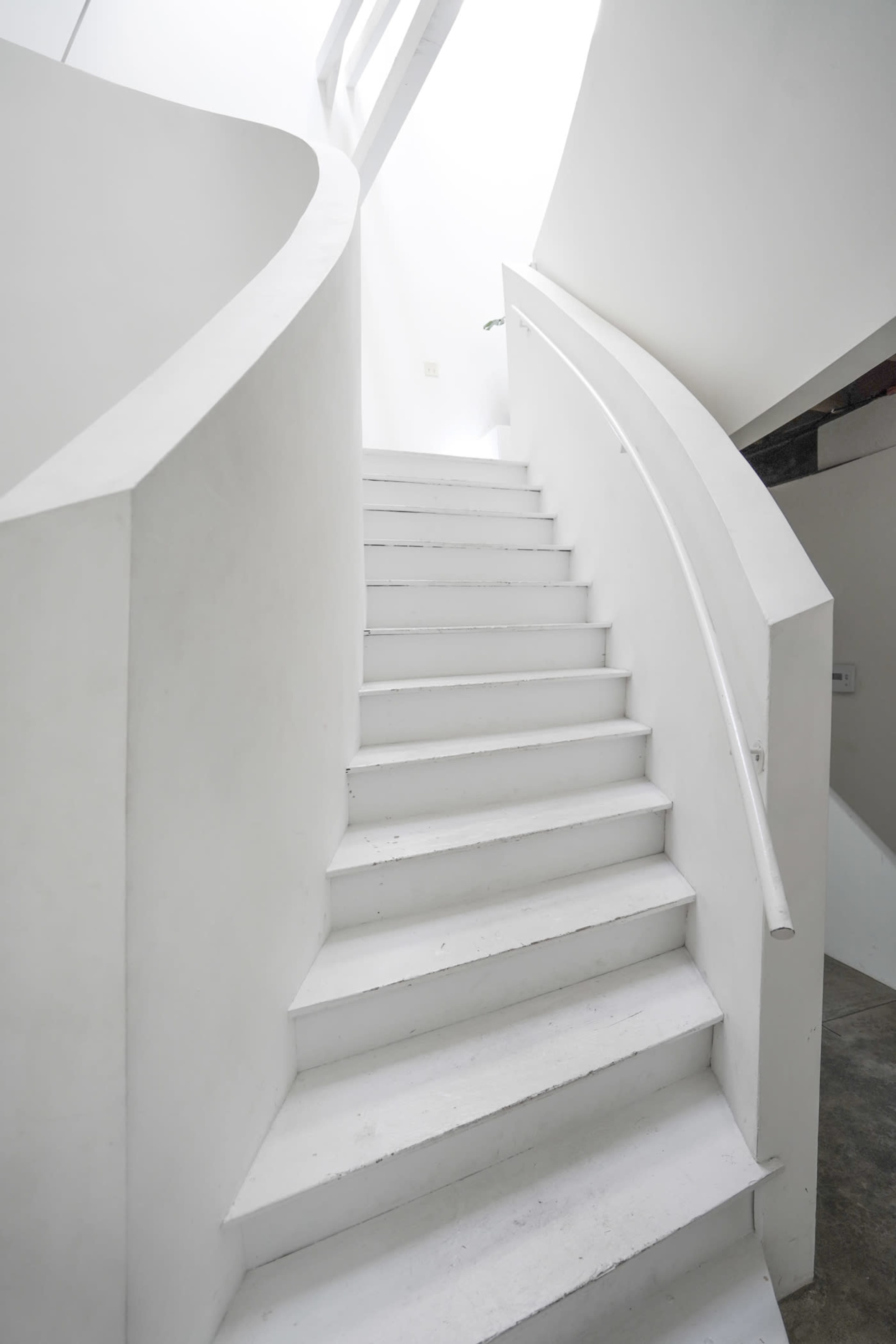 A spiral staircase with a smooth white railing ascends in a brightly lit, minimalist interior.