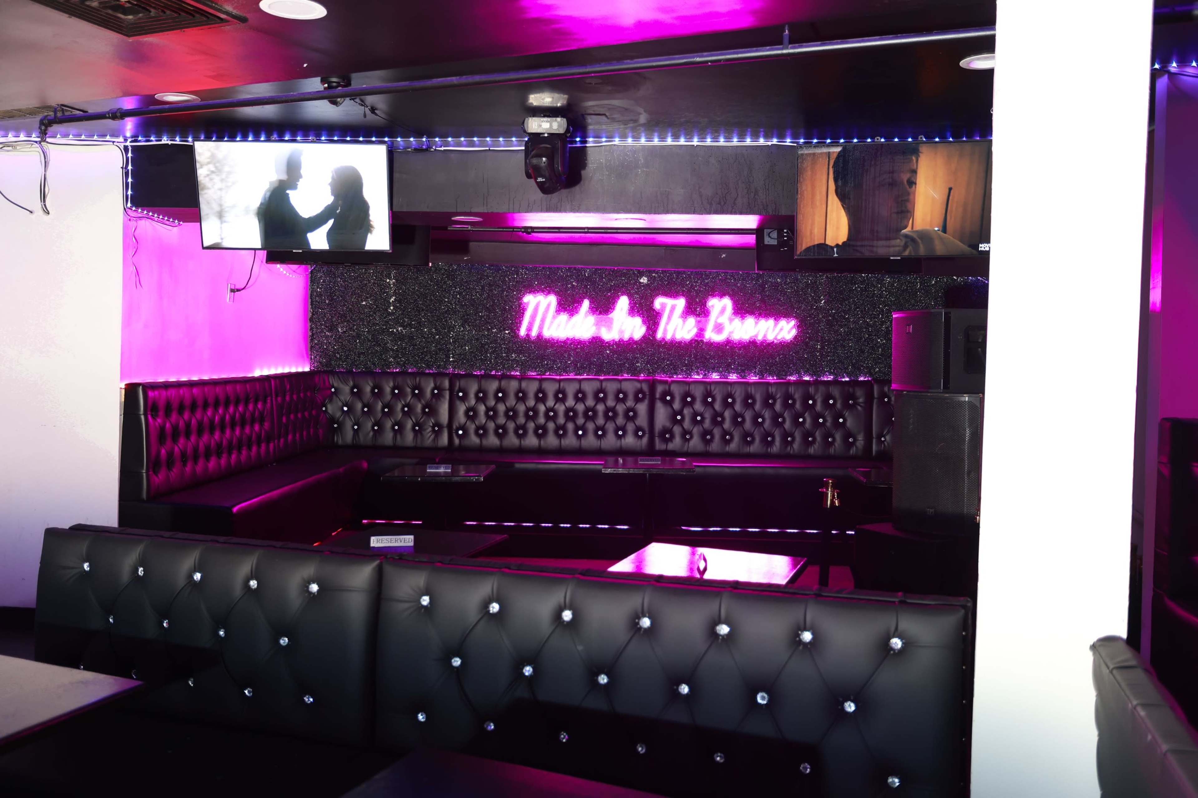 The image shows a dimly lit lounge area featuring black leather seating, LED lights, and screens displaying videos.
