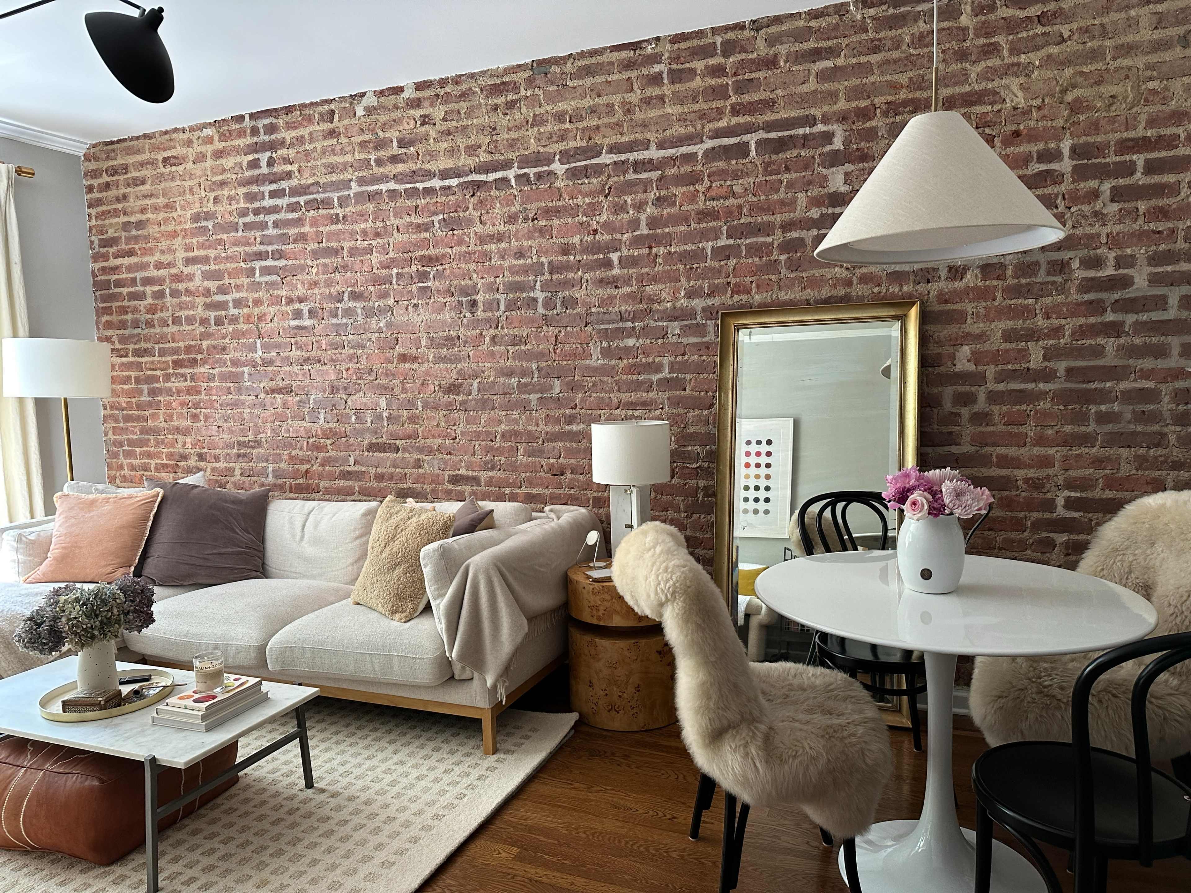 A cozy living space with a brick wall, a neutral sofa, a circular dining table with black chairs, and a decorative mirror.