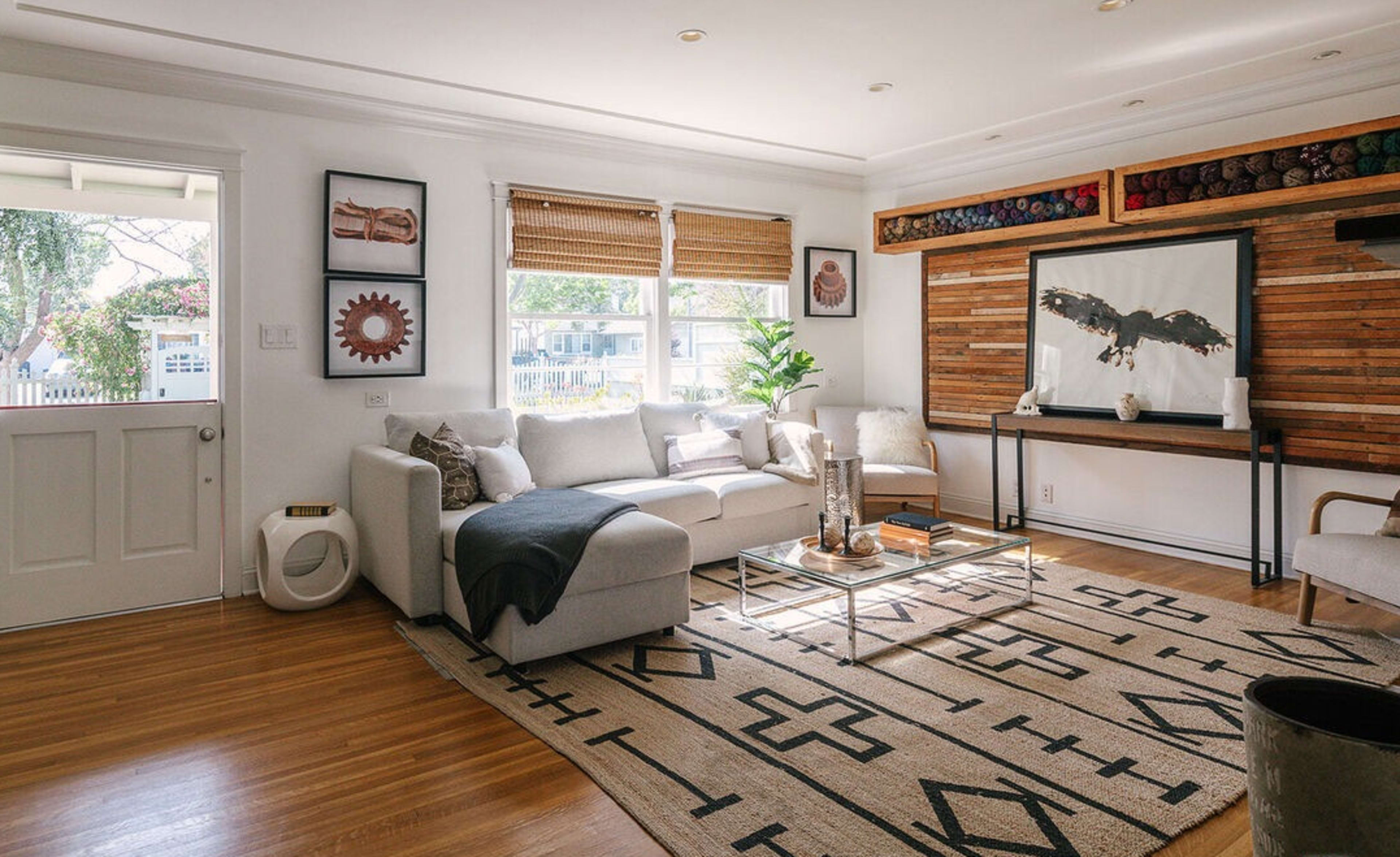 The image shows a bright living room with a sectional sofa, wooden accent wall, coffee table, and a door leading to the outside.
