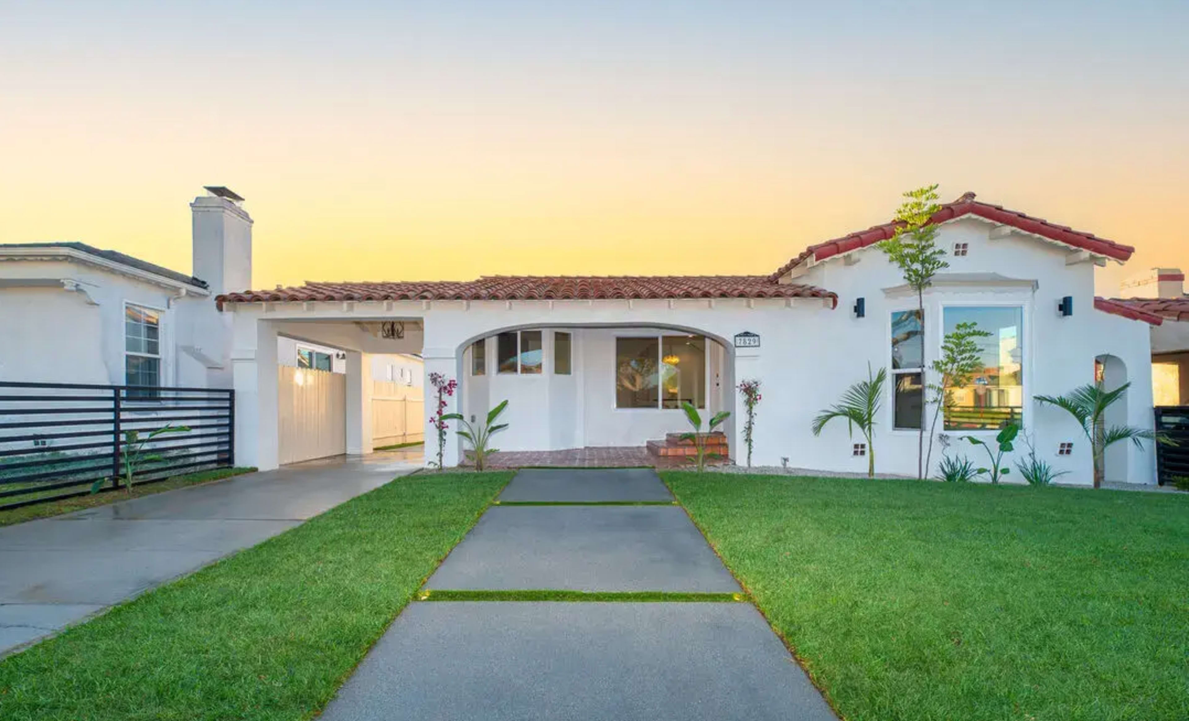 A white house with a red tile roof is set against a sunset backdrop, surrounded by green grass and bordered by a paved walkway.