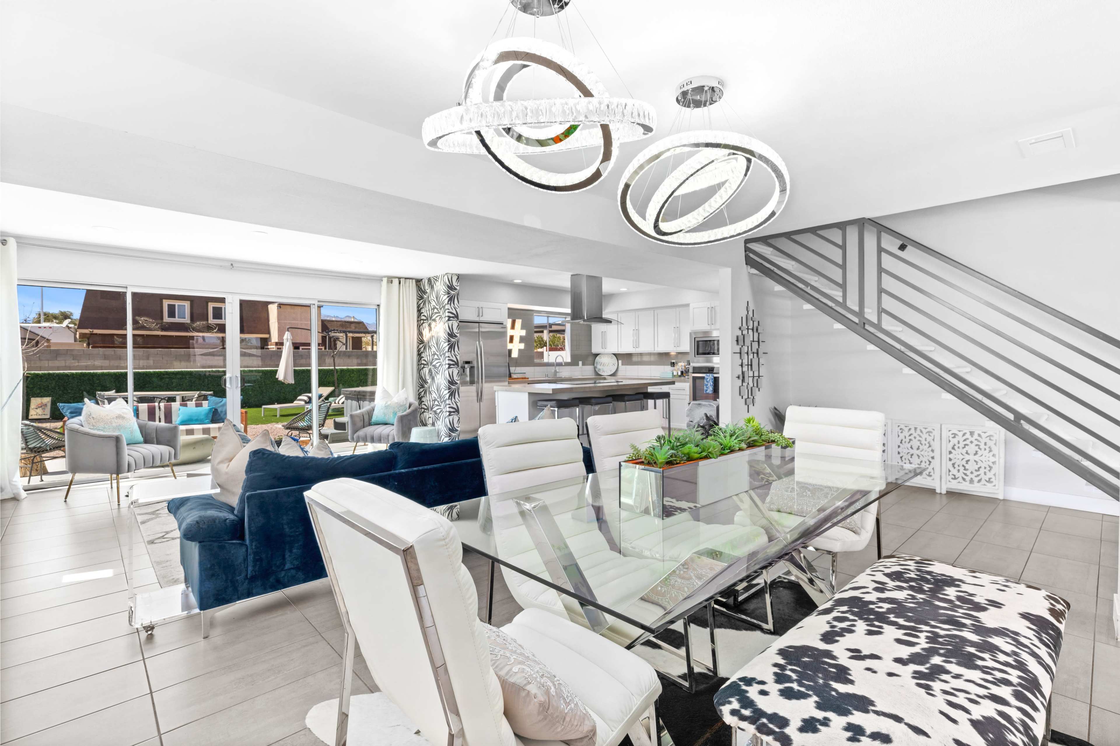 The image displays a modern dining area with a glass table surrounded by white chairs, adjacent to a stylish living space featuring a blue couch and sleek lighting fixtures.