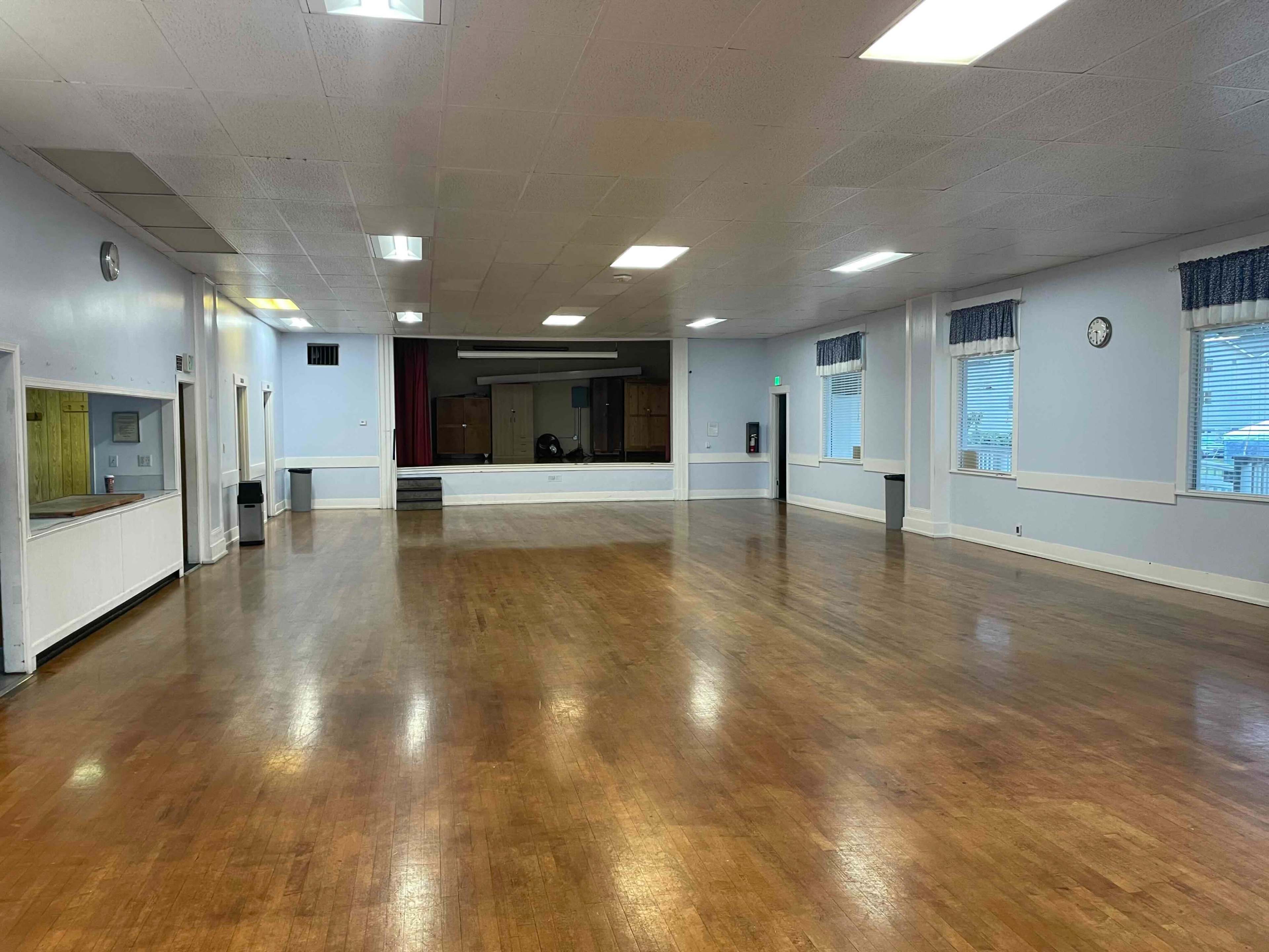 A large, empty hall with polished wooden floors, white walls, and a stage at the far end.