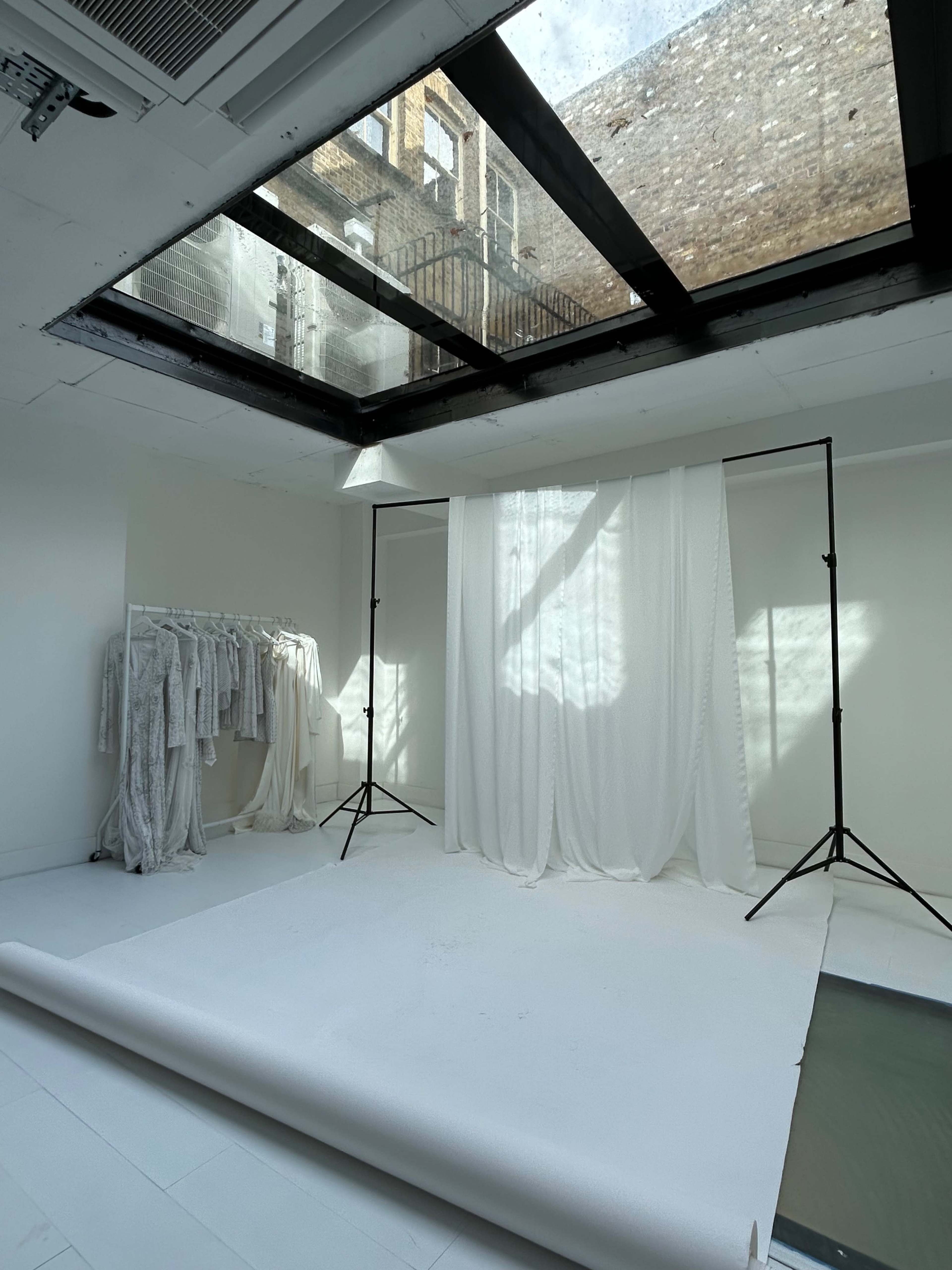 Abright white photo studio with a skylight, featuring a backdrop and hanging garments on a rack.