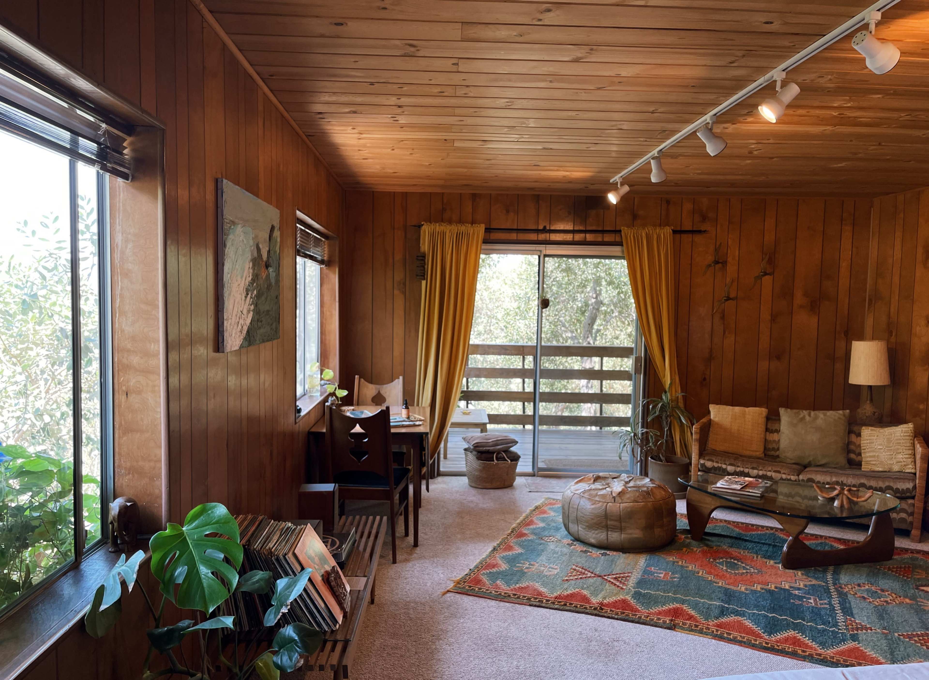 A cozy living room features wooden paneling, large windows with yellow curtains, and a combination of a seating area and a small dining space.