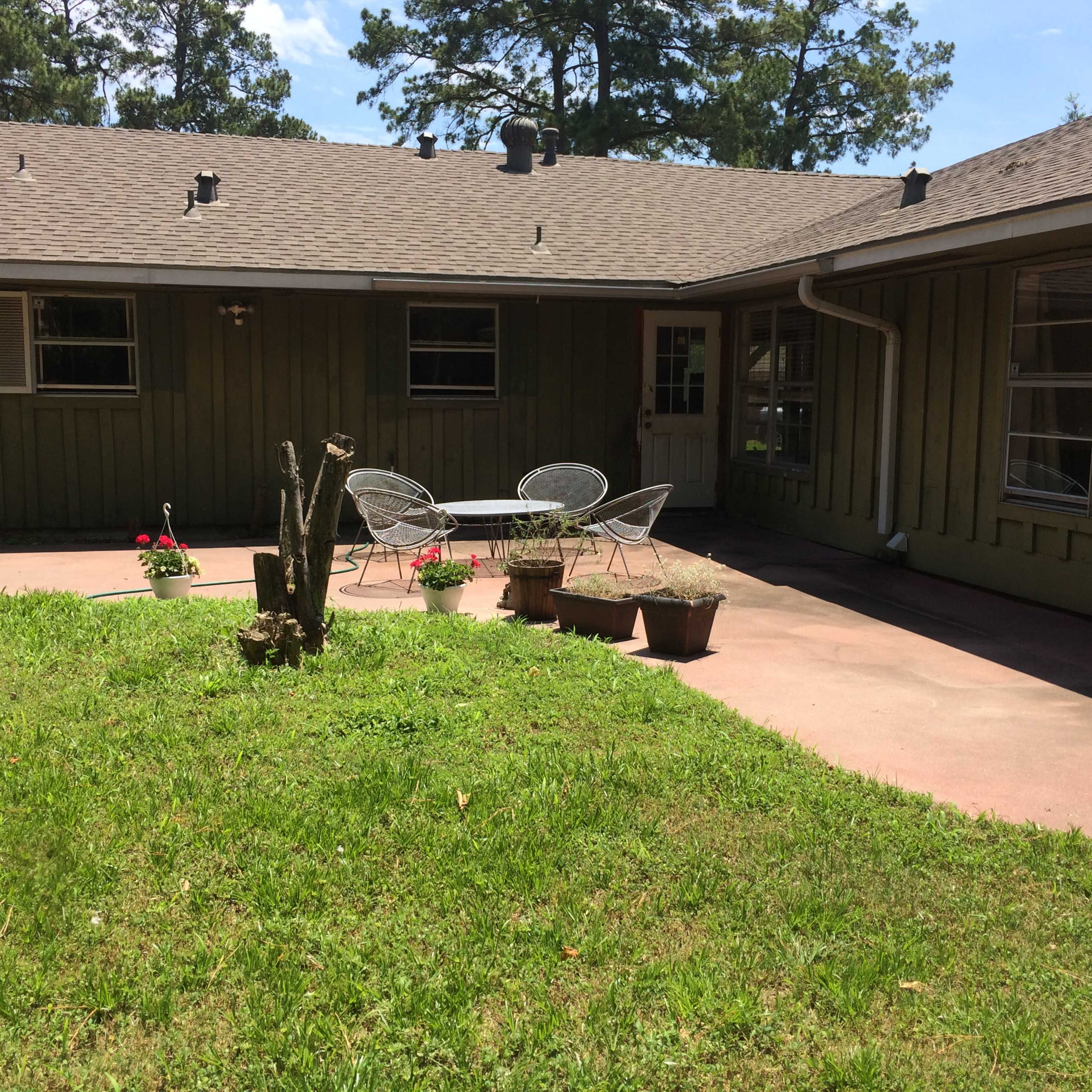 A paved patio with metal chairs and flower pots is surrounded by a grassy lawn next to a single-story house.