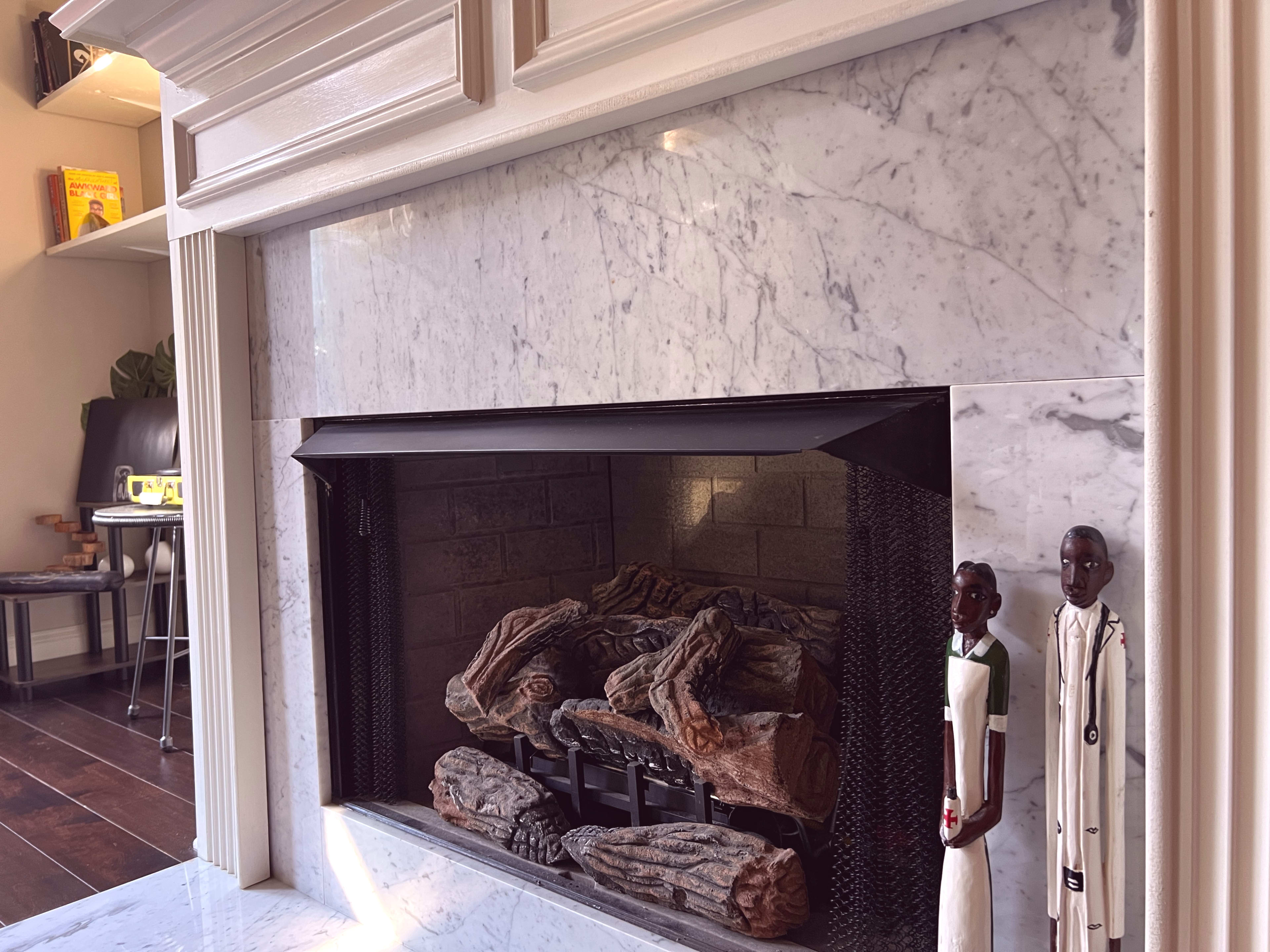 A modern fireplace with a marble surround, featuring decorative logs and two wooden figurines standing beside it.