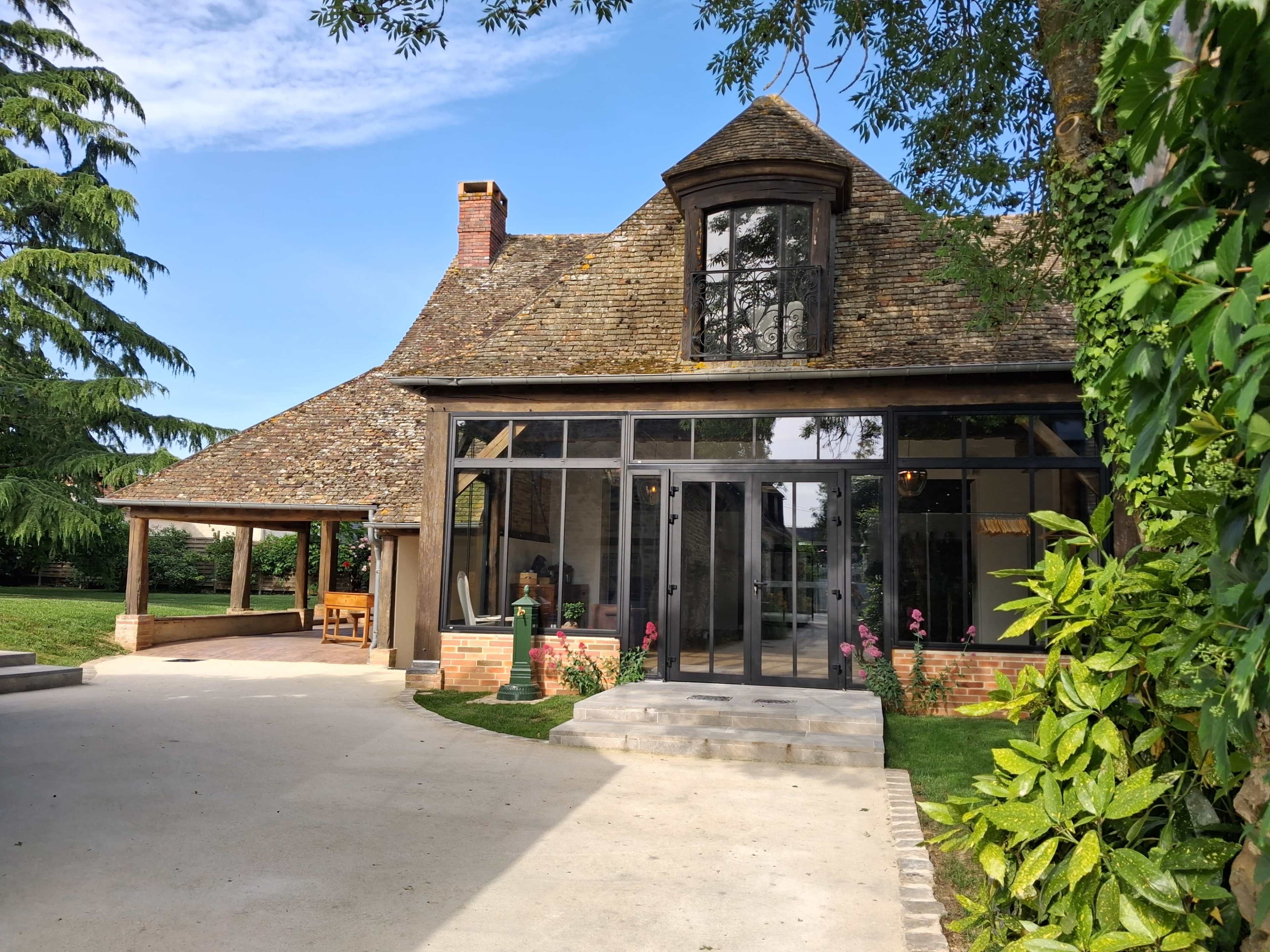 The image shows a house with a steeply pitched roof and large glass windows, surrounded by greenery and a patio area.