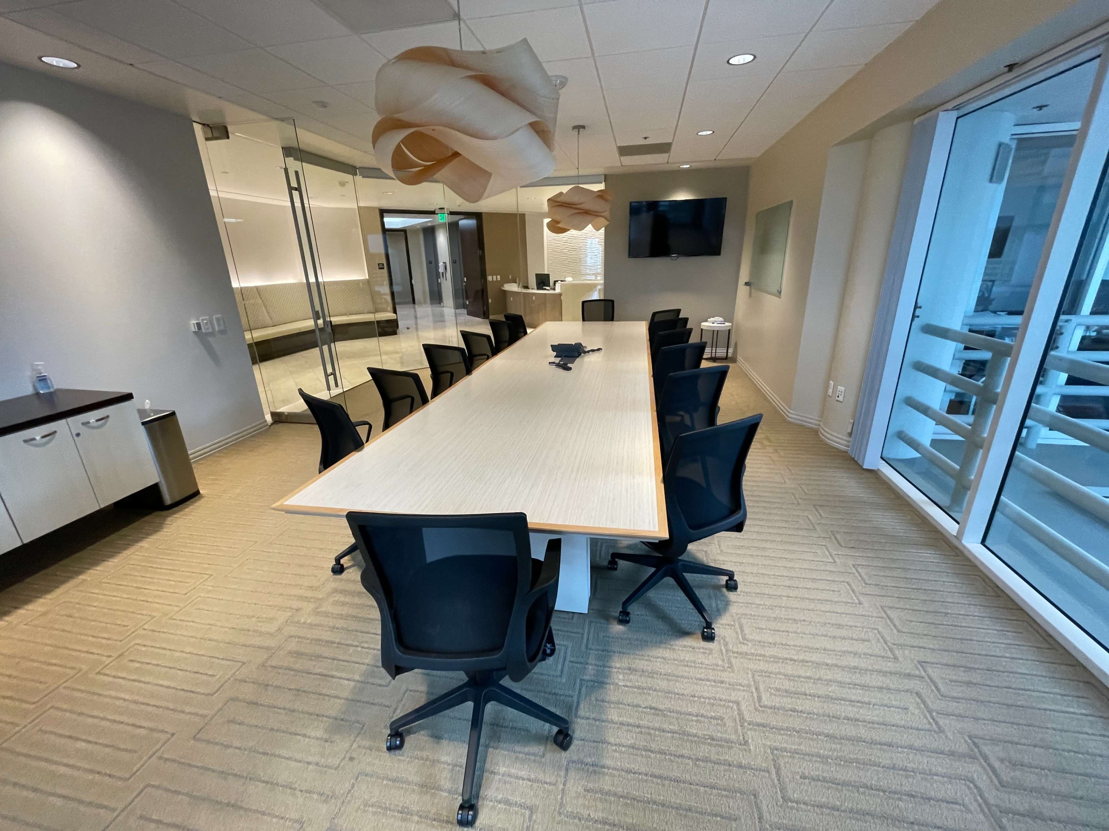 A modern conference room features a long table surrounded by black swivel chairs, with glass walls and overhead lighting.