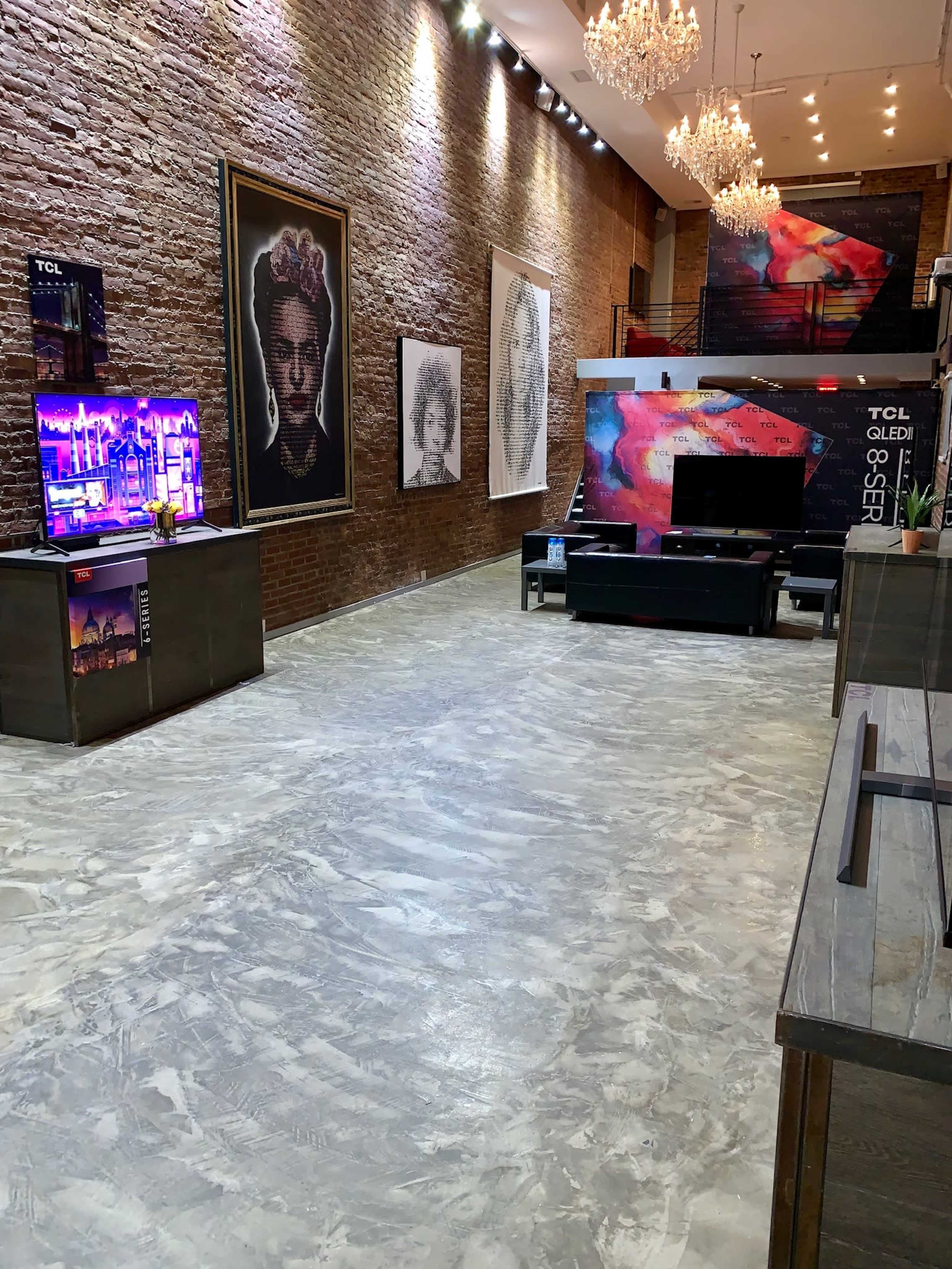 The image shows a modern showroom with a polished concrete floor, featuring various large artworks on the brick walls and multiple television displays.