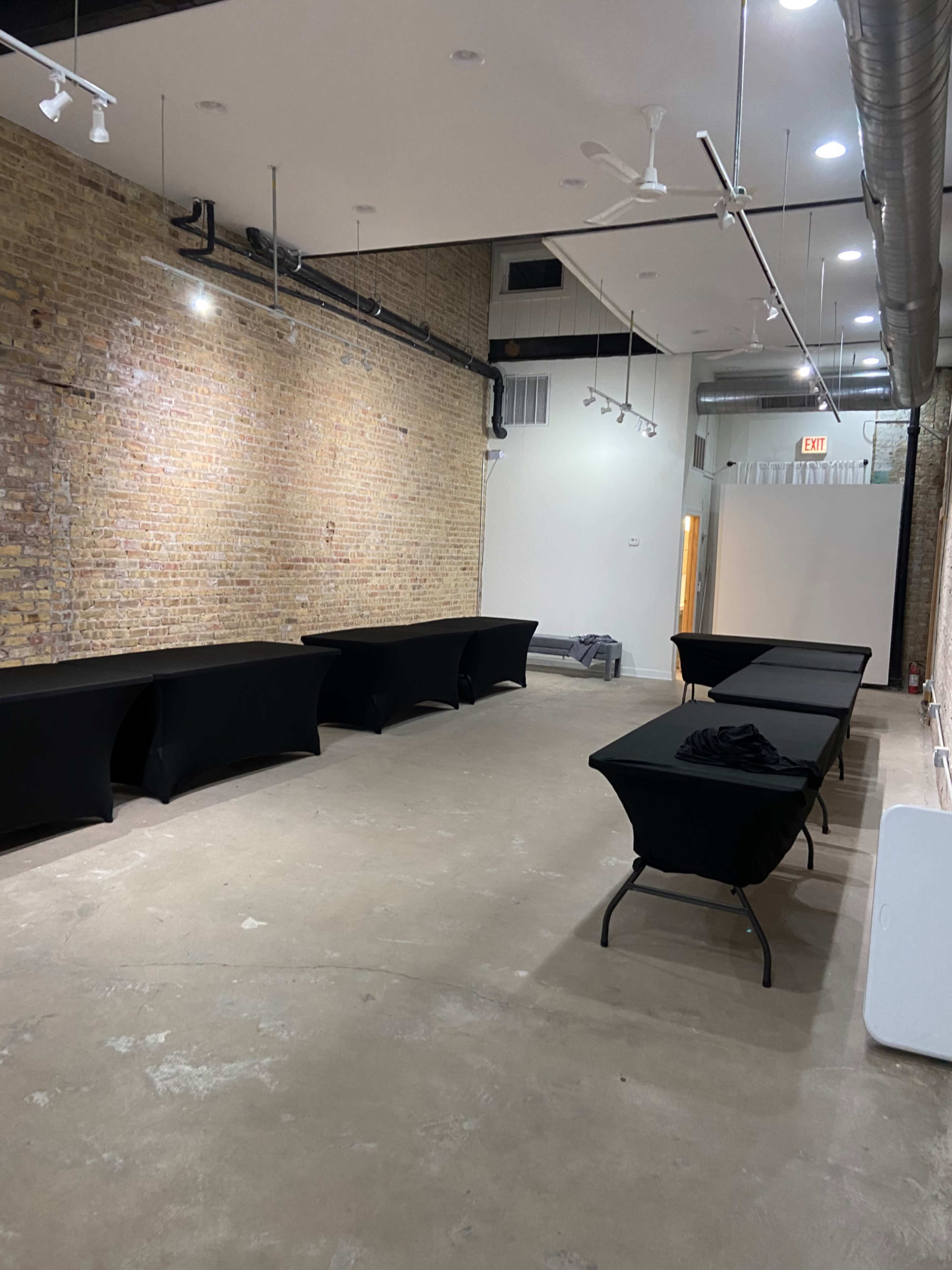 The image shows an empty room with exposed brick walls and several black tables arranged in a row.