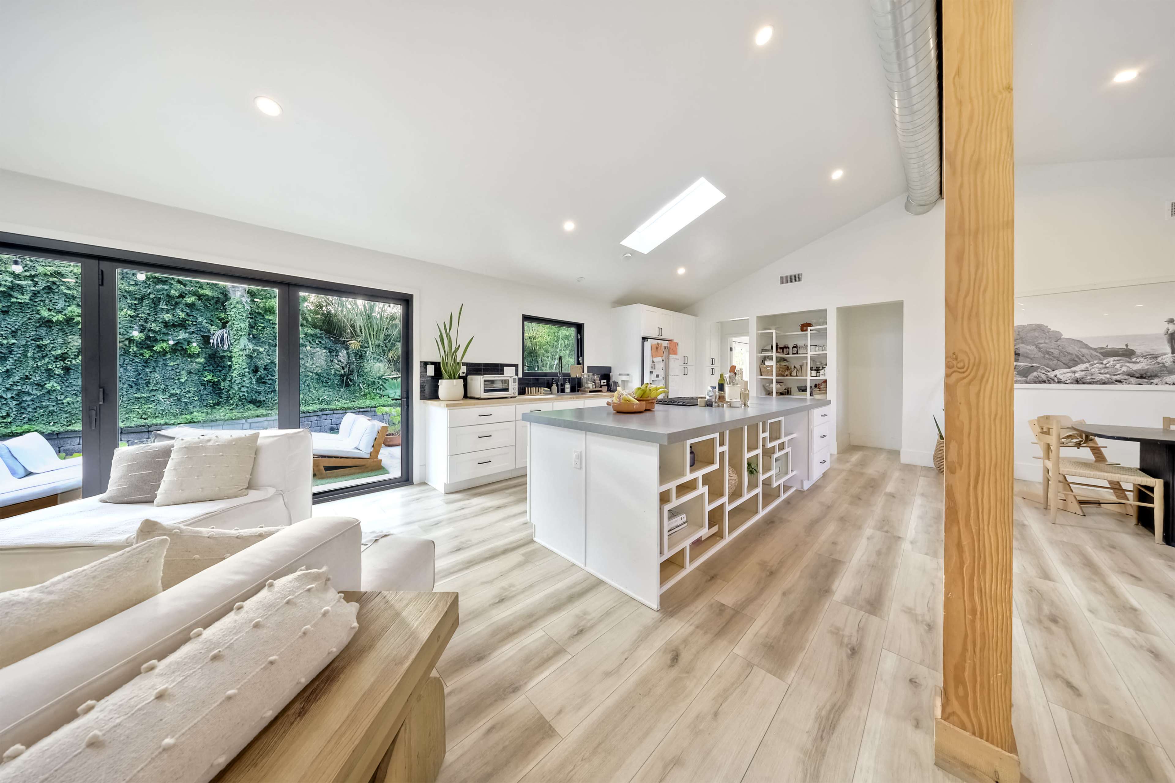 A modern kitchen and living area feature an open layout with wooden flooring, a central island, and large windows overlooking a garden.